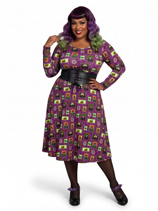 Plus-size model with a purple and lime green ombre Bettie Page hairstyle posing in a 1950s purple gothabilly swing dress featuring spooky monster frames and pumpkins. The long-sleeve, fit-and-flare midi dress is styled with a black corset belt, showcasing a flattering, inclusive curvy fit against a white background.