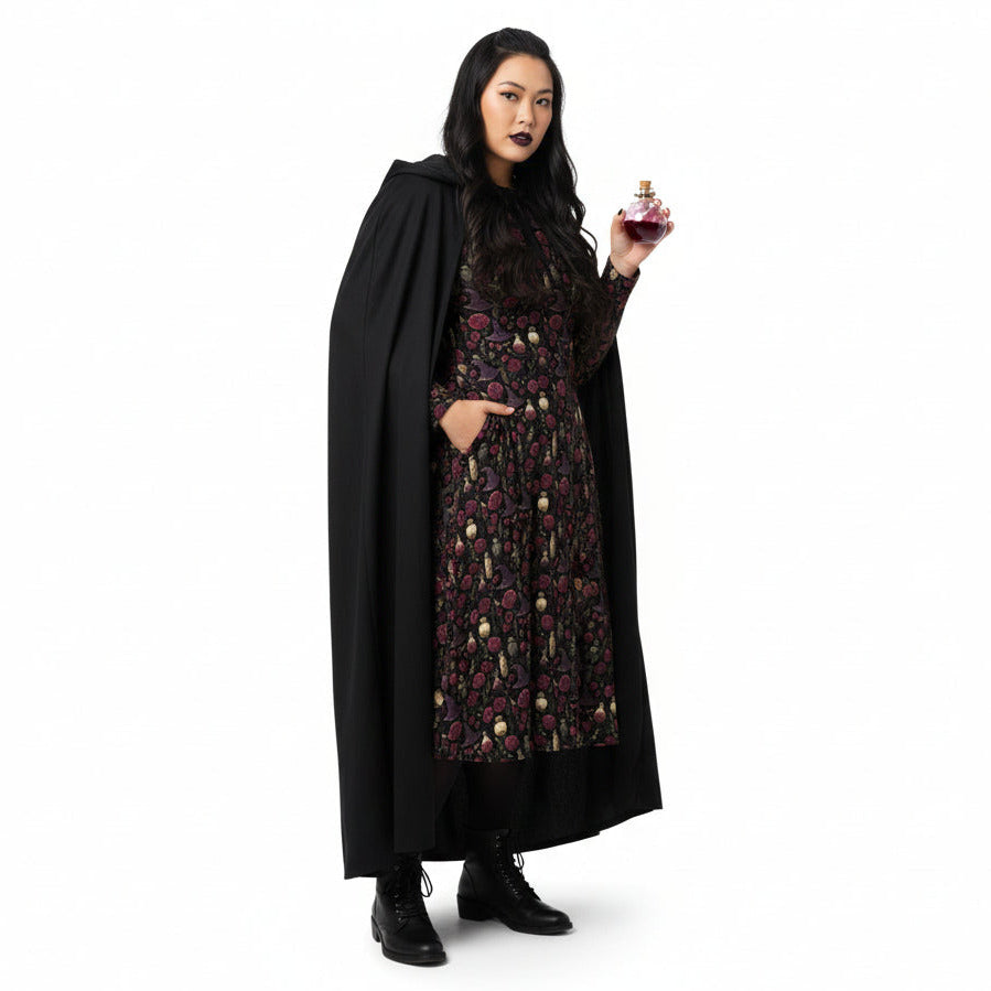 Woman wearing a black cape and patterned dress holding a glass of red wine on a white background