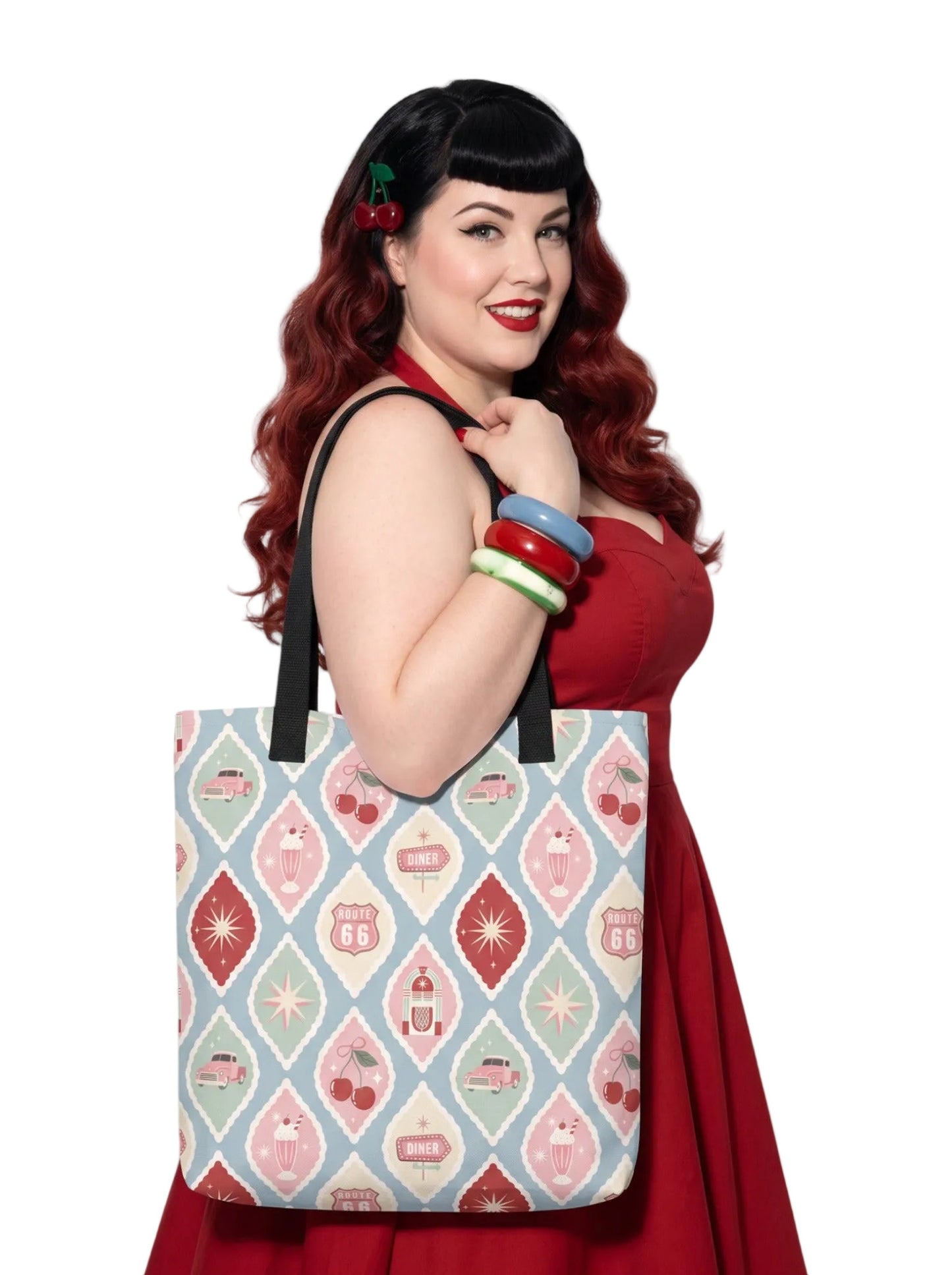 A woman holding a 1950s diner retro tote bag with pastel pinup pattern featuring cherries, milkshakes, and jukeboxes on a light blue background with black straps.