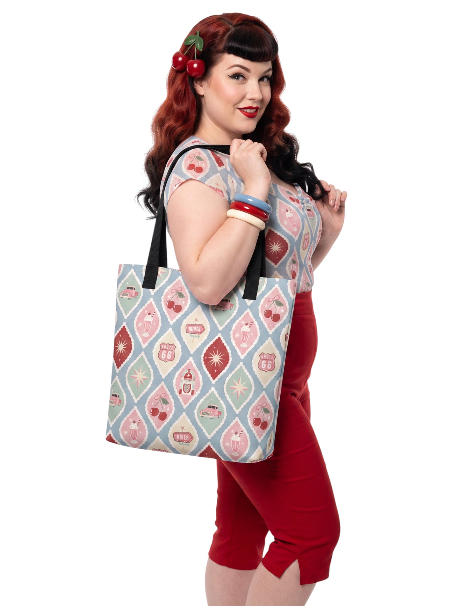 A woman holding a 1950s diner retro tote bag with pastel pinup pattern featuring cherries, milkshakes, and jukeboxes on a light blue background with black straps.