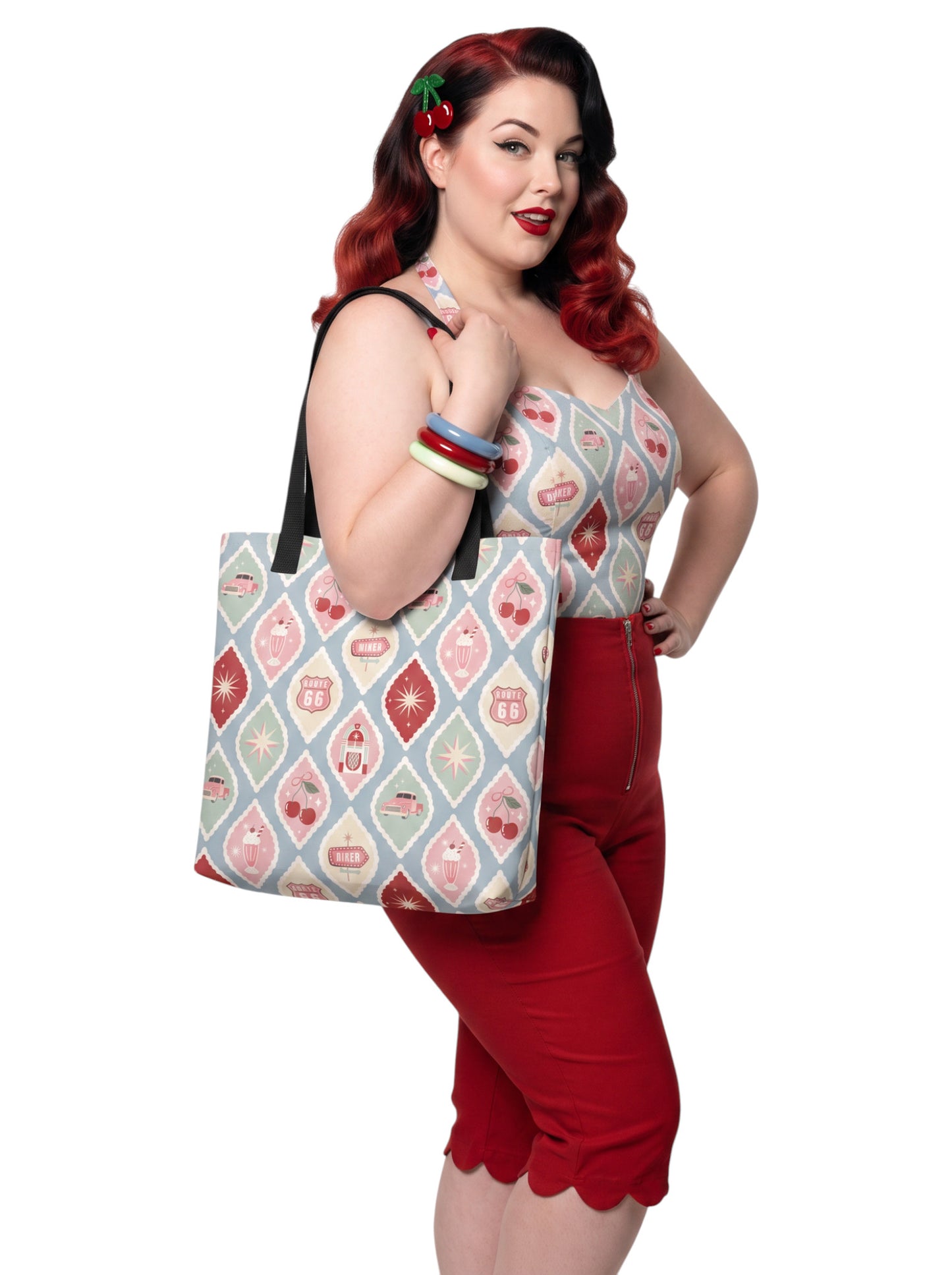 A woman holding a 1950s diner retro tote bag with pastel pinup pattern featuring cherries, milkshakes, and jukeboxes on a light blue background with black straps.