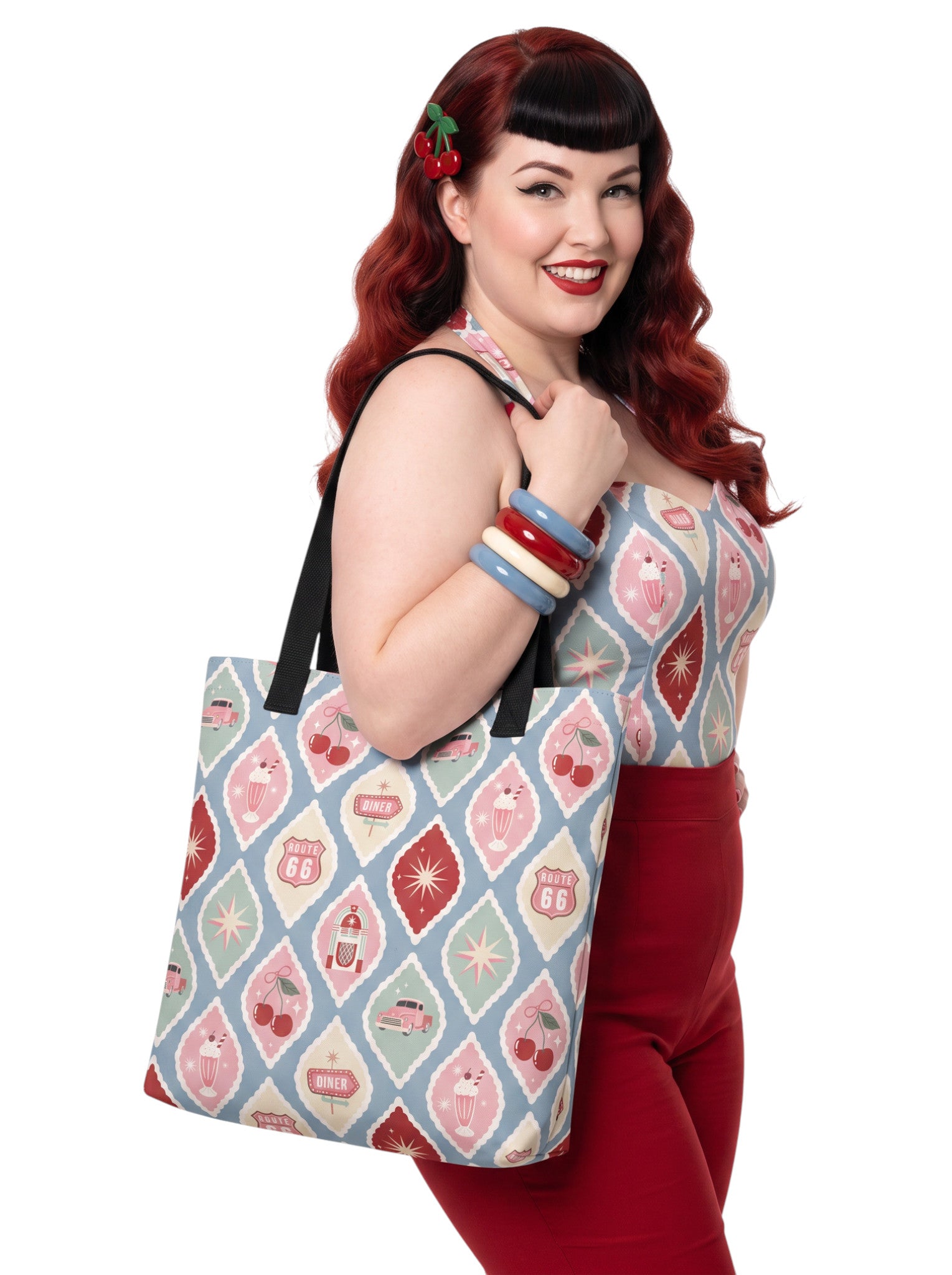A woman holding a 1950s diner retro tote bag with pastel pinup pattern featuring cherries, milkshakes, and jukeboxes on a light blue background with black straps.