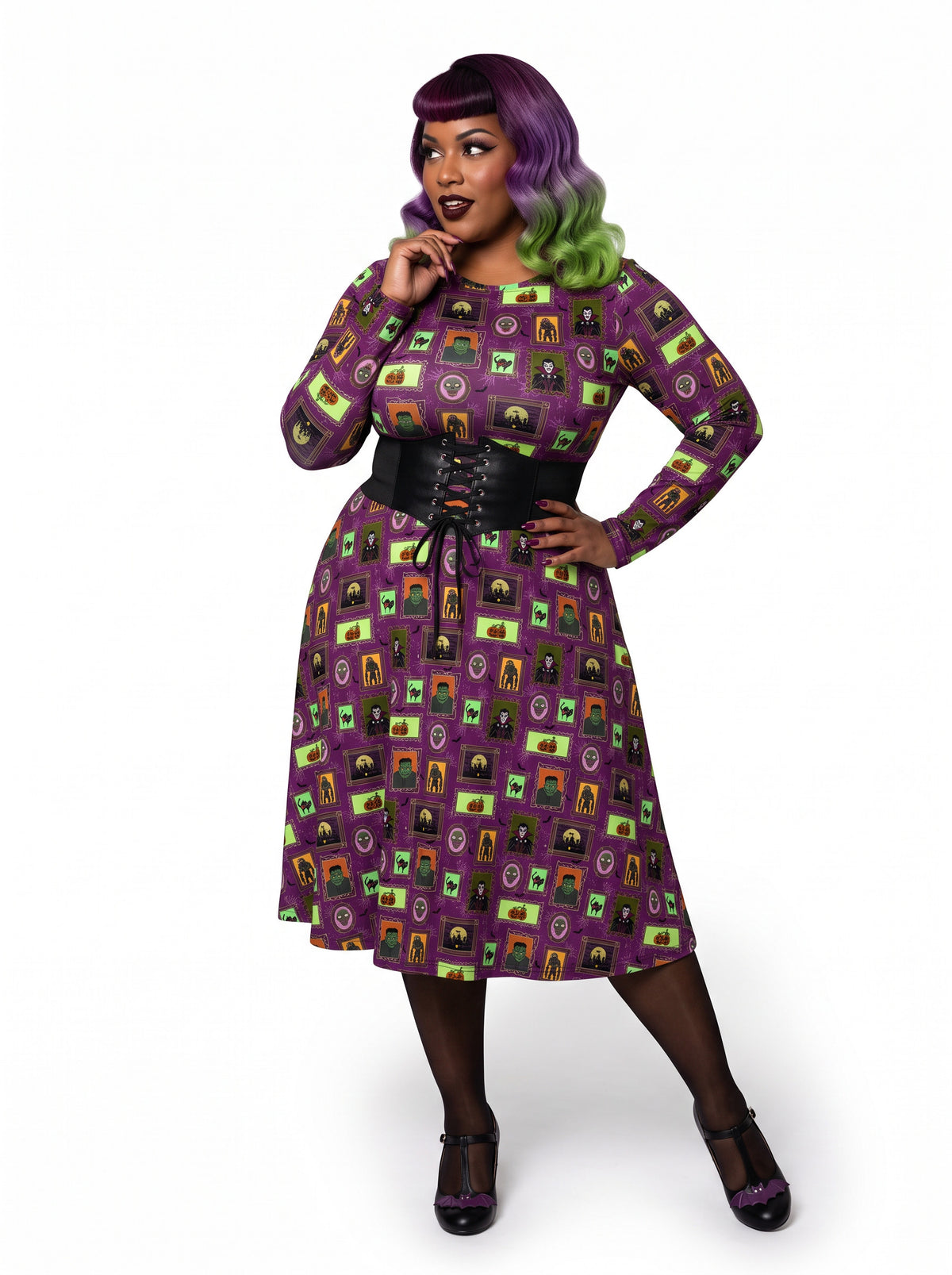 Plus-size model with a purple and lime green ombre Bettie Page hairstyle posing in a 1950s purple gothabilly swing dress featuring spooky monster frames and pumpkins. The long-sleeve, fit-and-flare midi dress is styled with a black corset belt, showcasing a flattering, inclusive curvy fit against a white background.