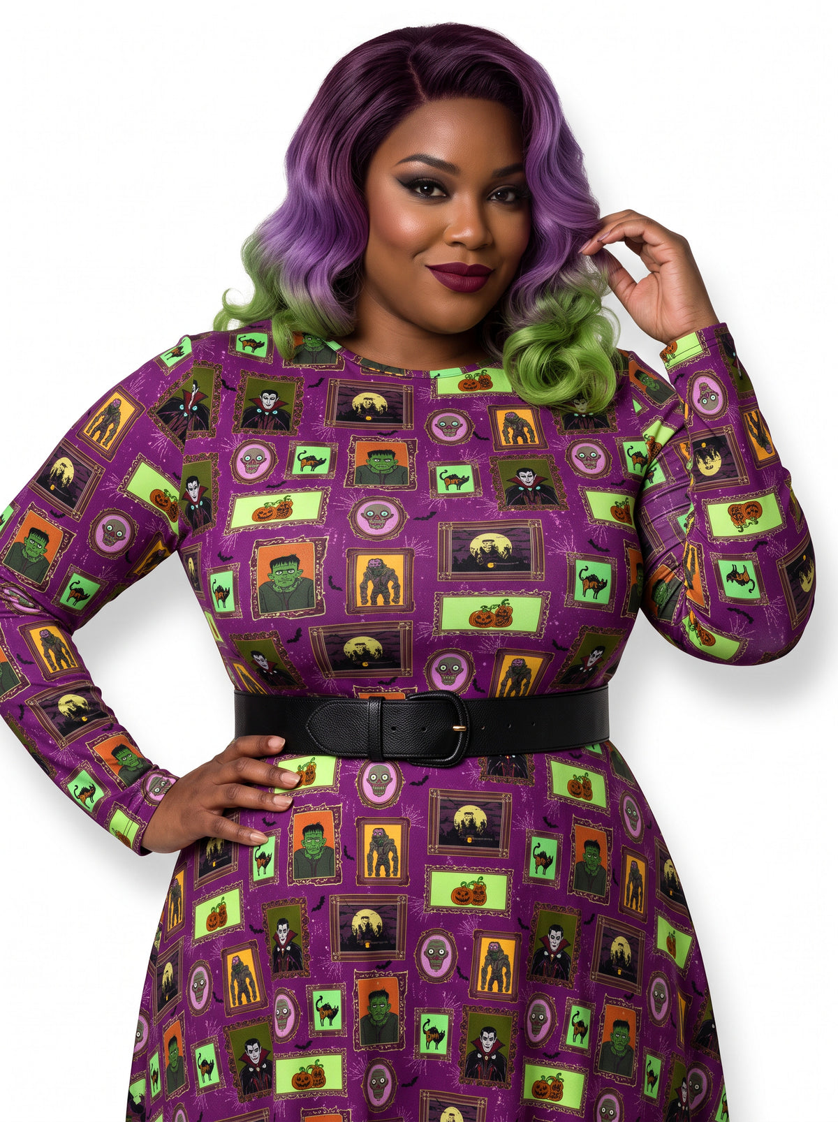 Plus-size model with a purple and lime green ombre Bettie Page hairstyle posing in a 1950s purple gothabilly swing dress featuring spooky monster frames and pumpkins. The long-sleeve, fit-and-flare midi dress is styled with a black corset belt, showcasing a flattering, inclusive curvy fit against a white background.