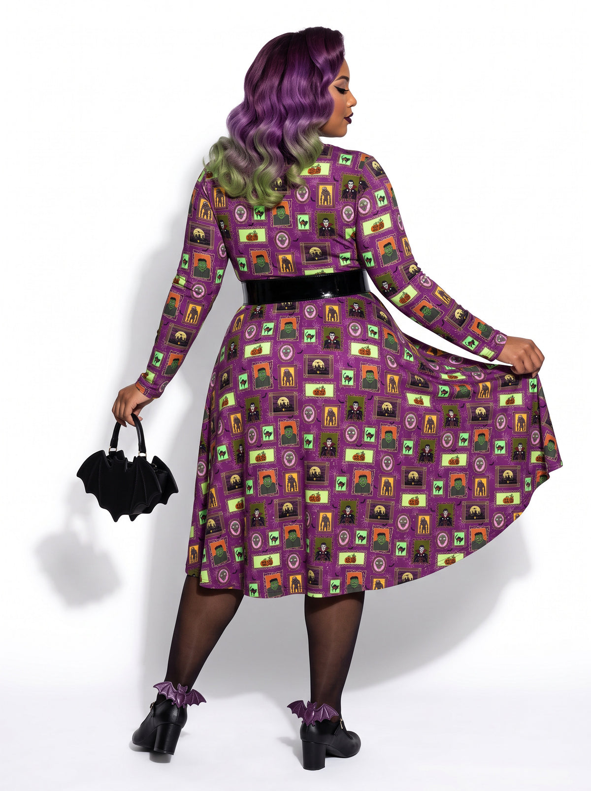 Plus-size model with a purple and lime green ombre Bettie Page hairstyle posing in a 1950s purple gothabilly swing dress featuring spooky monster frames and pumpkins. The long-sleeve, fit-and-flare midi dress is styled with a black corset belt, showcasing a flattering, inclusive curvy fit against a white background.