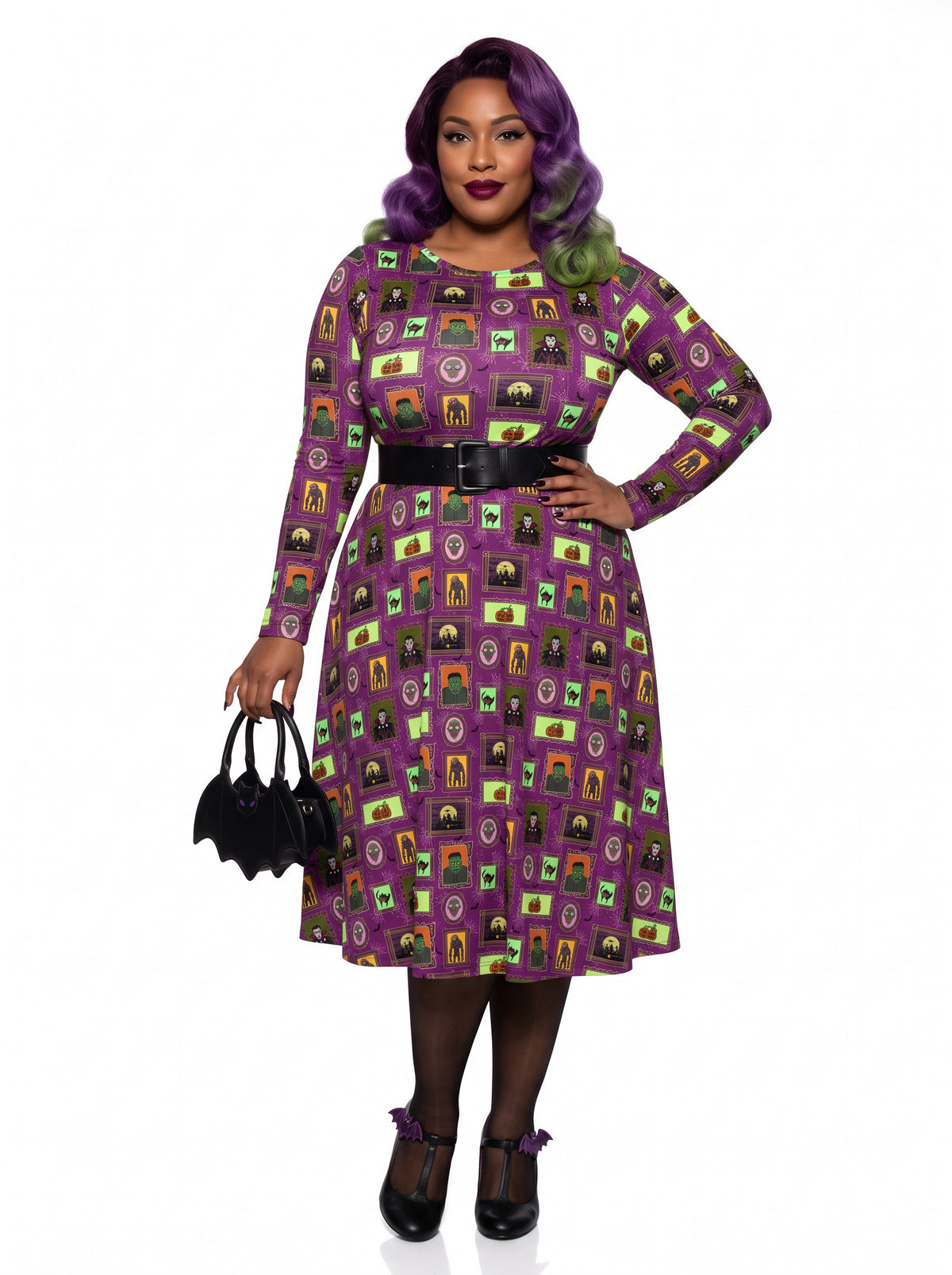 Plus-size model with a purple and lime green ombre Bettie Page hairstyle posing in a 1950s purple gothabilly swing dress featuring spooky monster frames and pumpkins. The long-sleeve, fit-and-flare midi dress is styled with a black corset belt, showcasing a flattering, inclusive curvy fit against a white background.