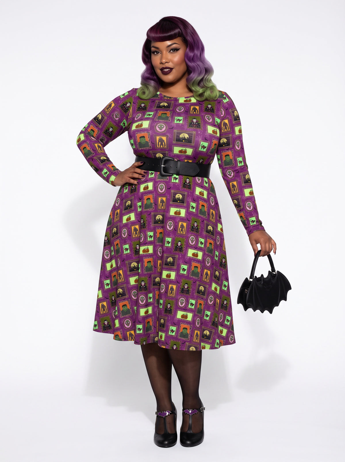 Plus-size model with a purple and lime green ombre Bettie Page hairstyle posing in a 1950s purple gothabilly swing dress featuring spooky monster frames and pumpkins. The long-sleeve, fit-and-flare midi dress is styled with a black corset belt, showcasing a flattering, inclusive curvy fit against a white background.