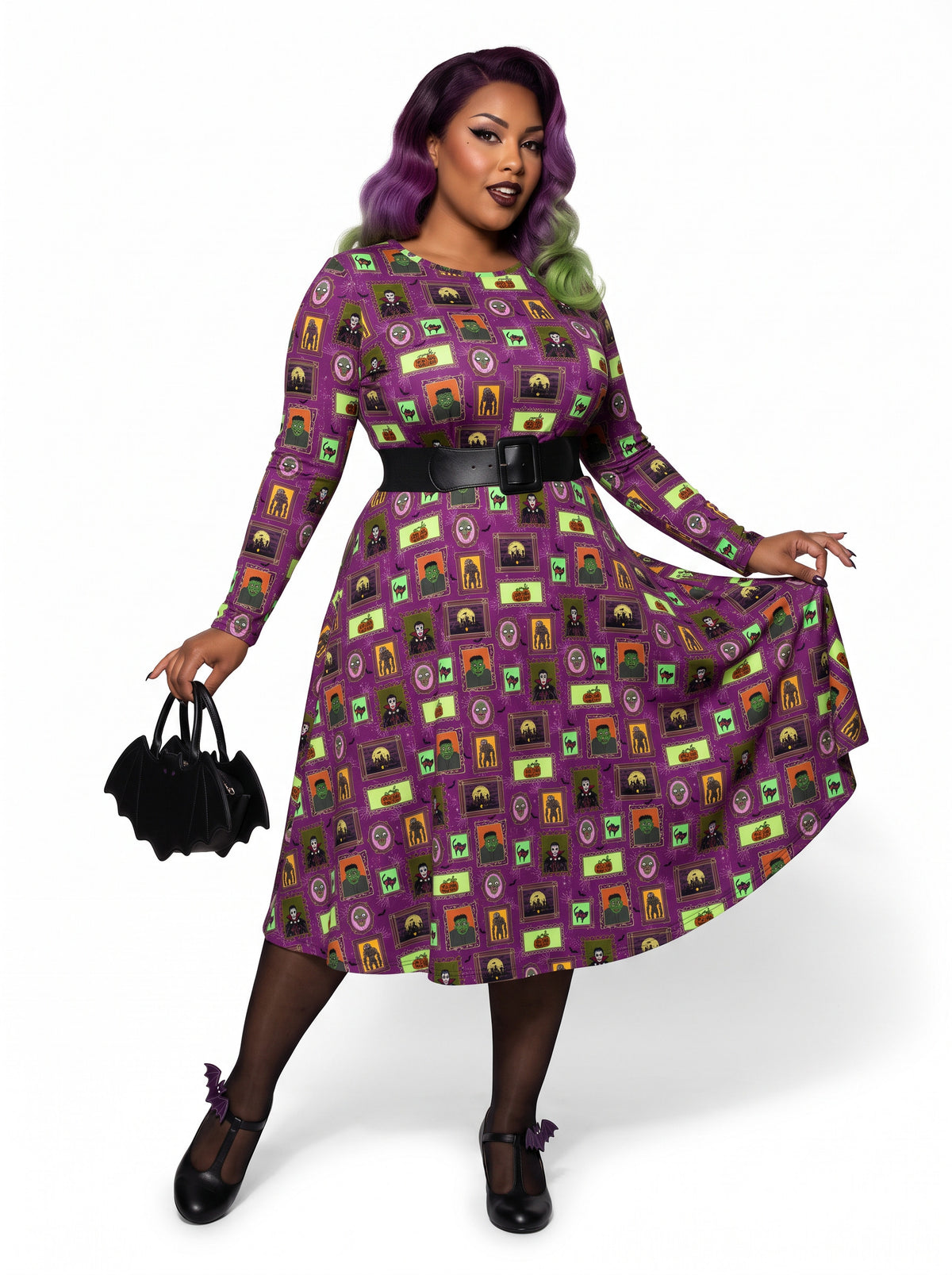 Plus-size model with a purple and lime green ombre Bettie Page hairstyle posing in a 1950s purple gothabilly swing dress featuring spooky monster frames and pumpkins. The long-sleeve, fit-and-flare midi dress is styled with a black corset belt, showcasing a flattering, inclusive curvy fit against a white background.