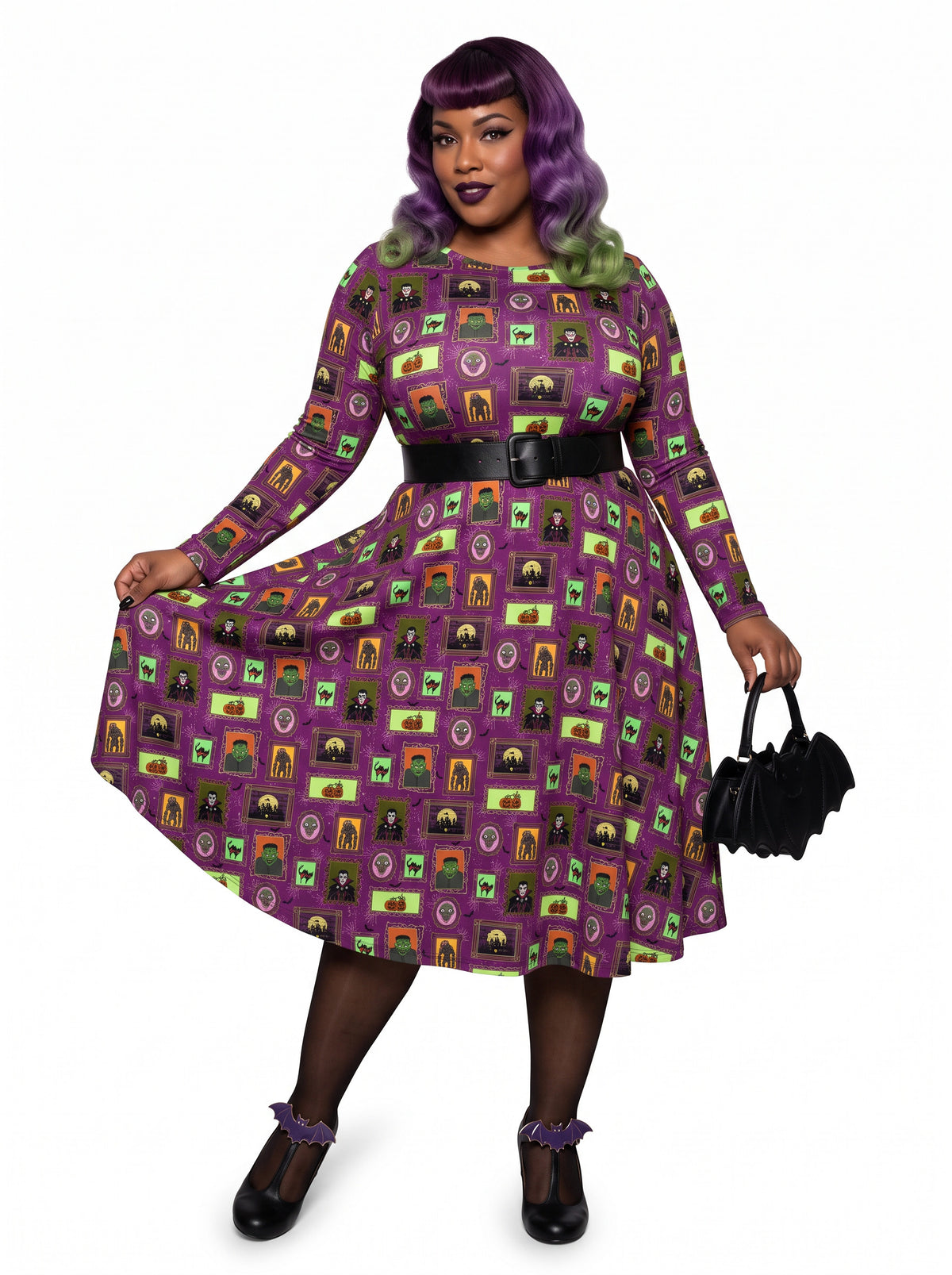Plus-size model with a purple and lime green ombre Bettie Page hairstyle posing in a 1950s purple gothabilly swing dress featuring spooky monster frames and pumpkins. The long-sleeve, fit-and-flare midi dress is styled with a black corset belt, showcasing a flattering, inclusive curvy fit against a white background.