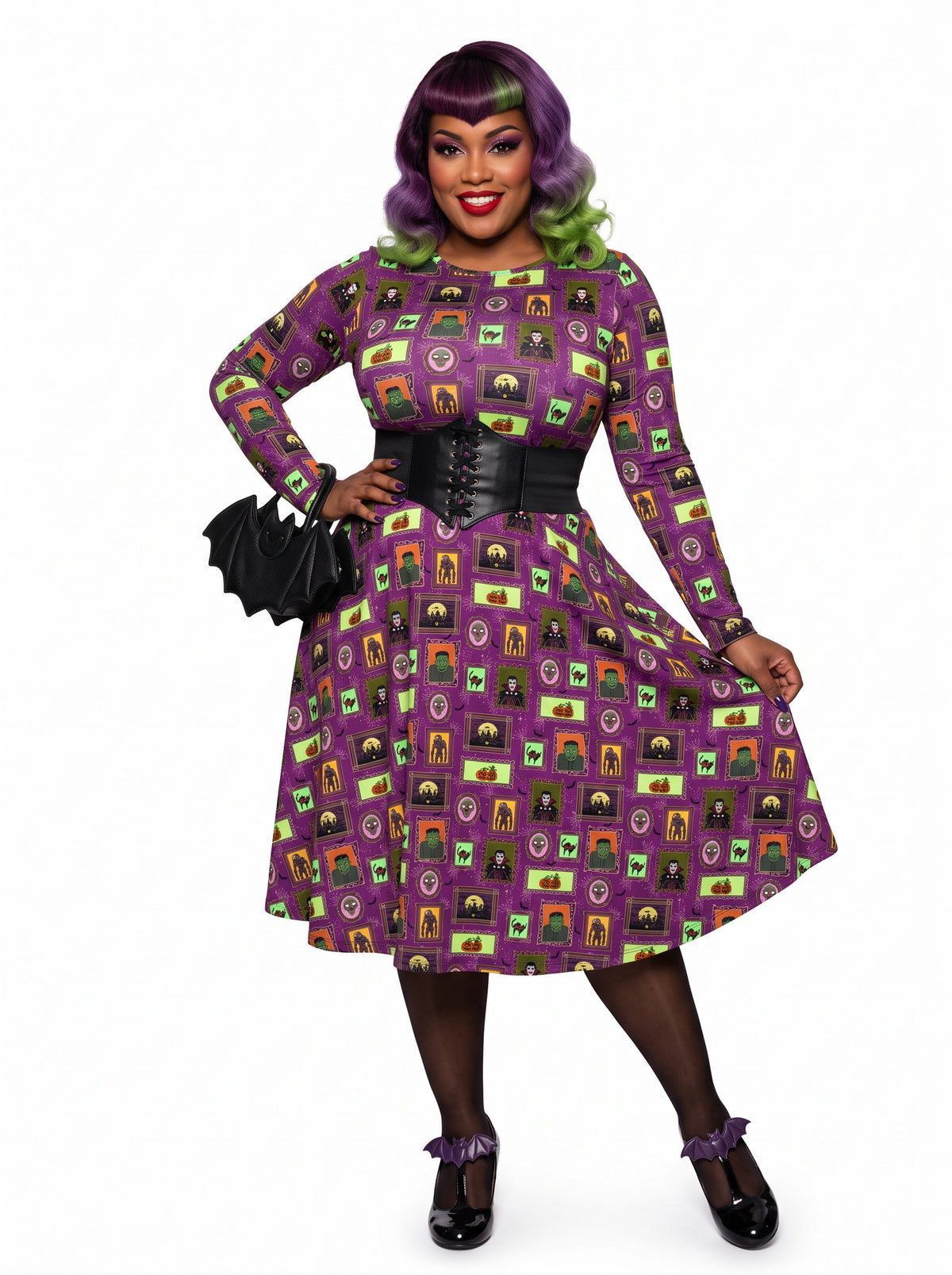 Plus-size model with a purple and lime green ombre Bettie Page hairstyle posing in a 1950s purple gothabilly swing dress featuring spooky monster frames and pumpkins. The long-sleeve, fit-and-flare midi dress is styled with a black corset belt, showcasing a flattering, inclusive curvy fit against a white background.