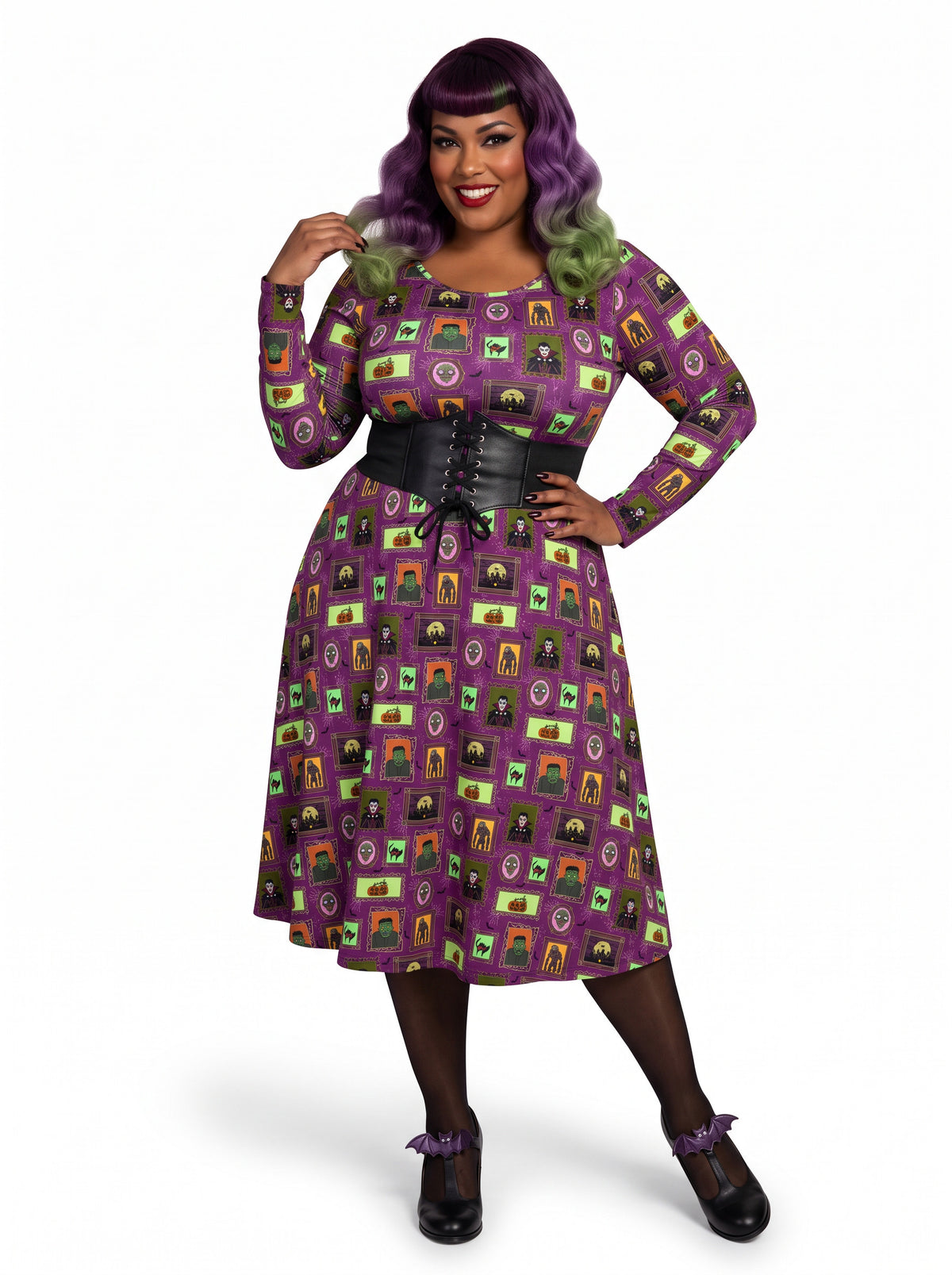Plus-size model with a purple and lime green ombre Bettie Page hairstyle posing in a 1950s purple gothabilly swing dress featuring spooky monster frames and pumpkins. The long-sleeve, fit-and-flare midi dress is styled with a black corset belt, showcasing a flattering, inclusive curvy fit against a white background.