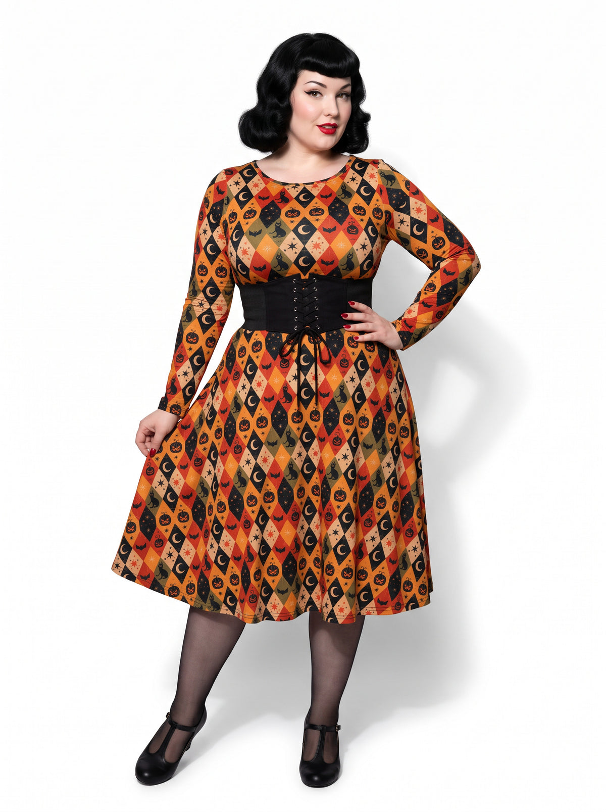 Plus-size model posing in a 1950s-style orange and black harlequin-patterned pinup midi swing dress featuring printed spooky pumpkins, bats, and crescent moons. The vintage-style dress shows a long-sleeve, fit-and-flare silhouette made from a smooth stretch fabric tailored for an inclusive, curvy fit.
