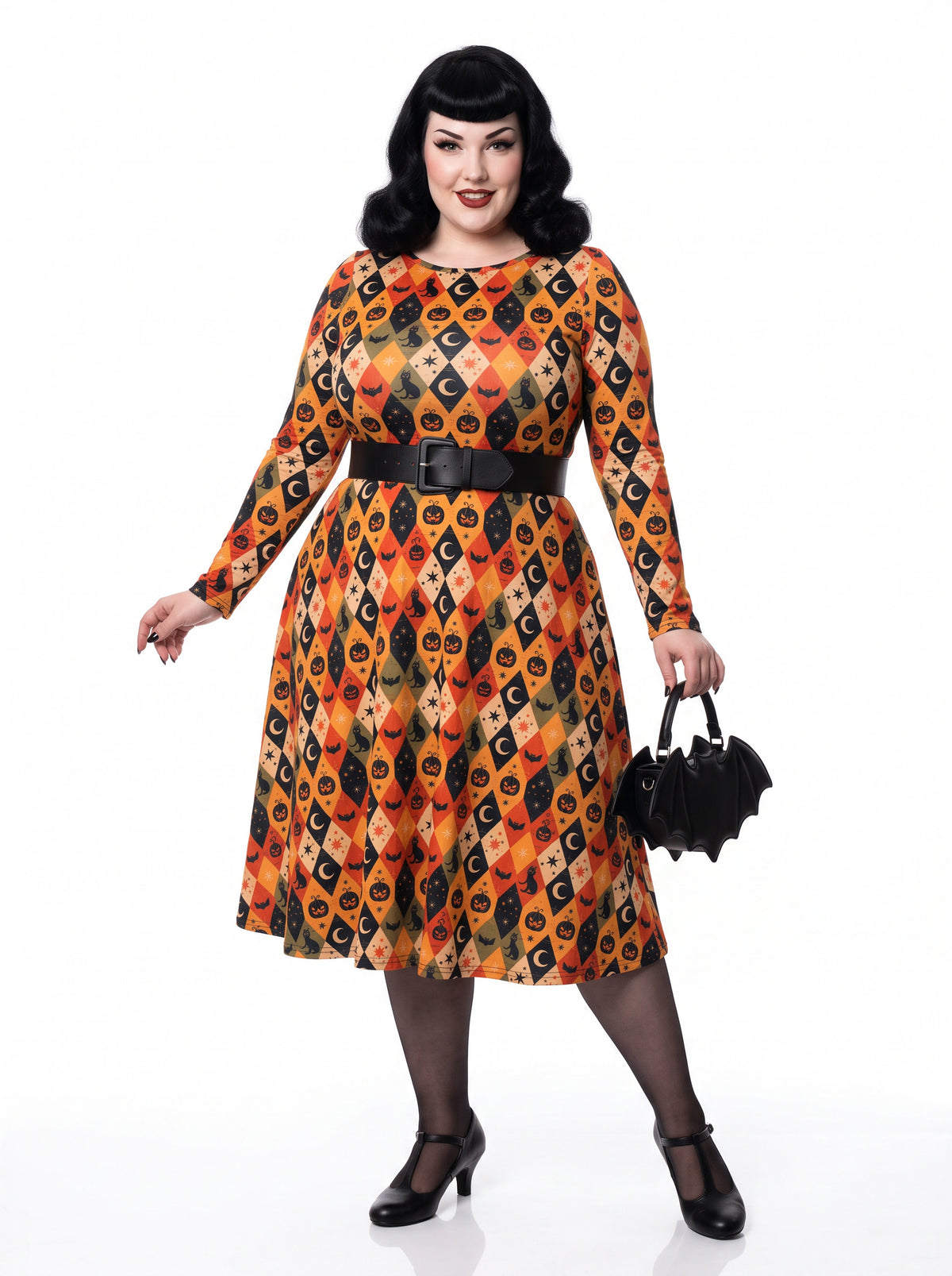 Plus-size model posing in a 1950s-style orange and black harlequin-patterned pinup midi swing dress featuring printed spooky pumpkins, bats, and crescent moons. The vintage-style dress shows a long-sleeve, fit-and-flare silhouette made from a smooth stretch fabric tailored for an inclusive, curvy fit.
