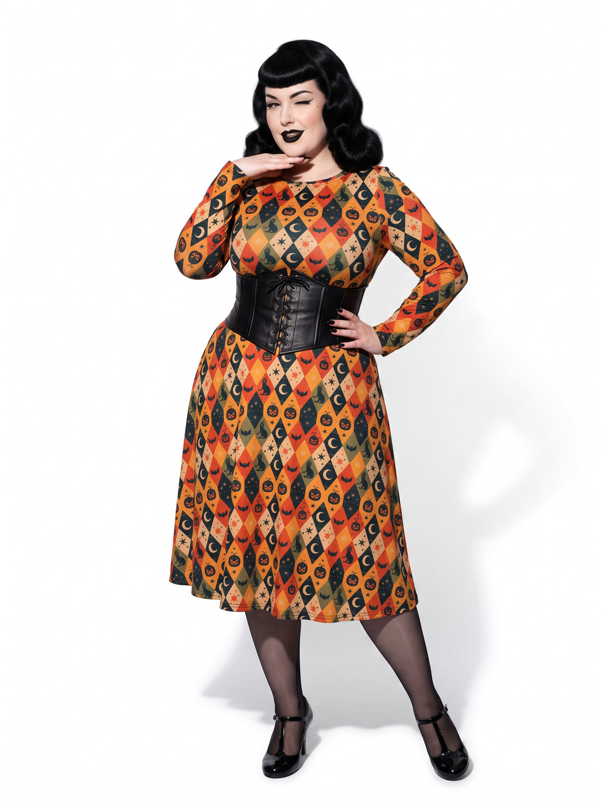 Plus-size model posing in a 1950s-style orange and black harlequin-patterned pinup midi swing dress featuring printed spooky pumpkins, bats, and crescent moons. The vintage-style dress shows a long-sleeve, fit-and-flare silhouette made from a smooth stretch fabric tailored for an inclusive, curvy fit.
