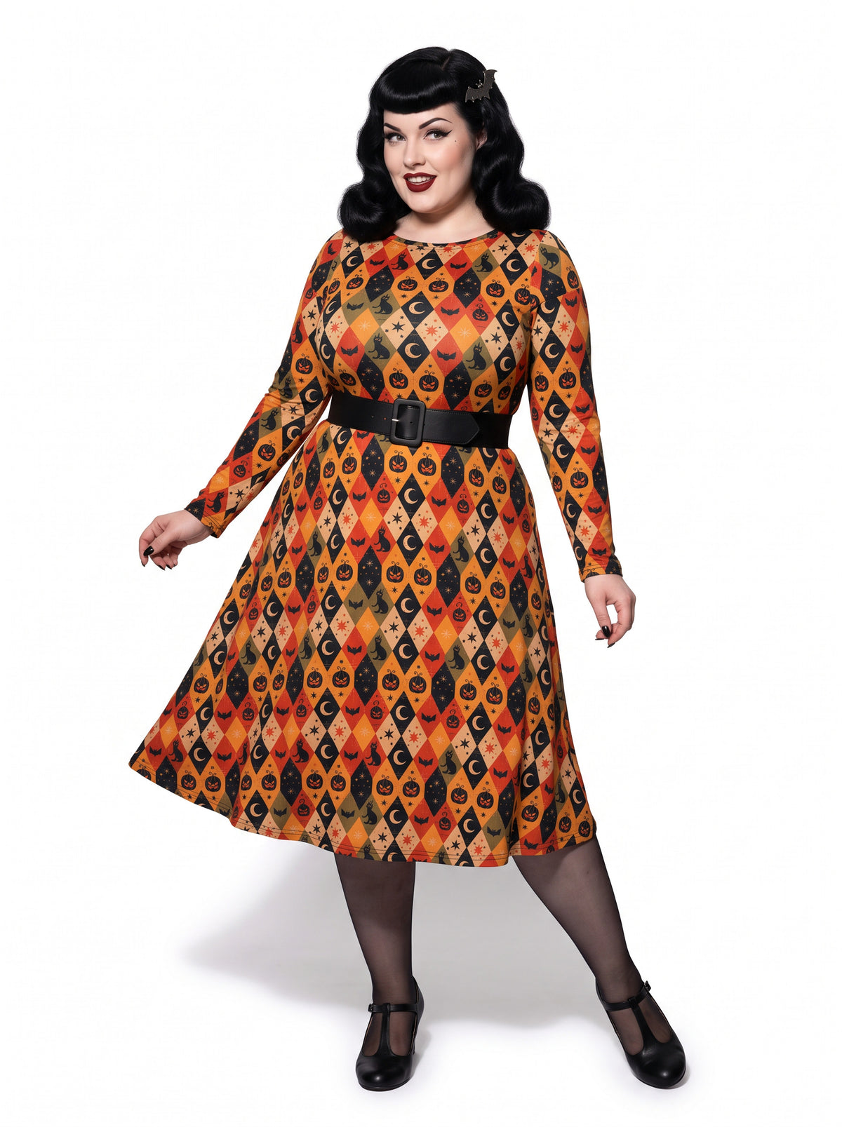 Plus-size model posing in a 1950s-style orange and black harlequin-patterned pinup midi swing dress featuring printed spooky pumpkins, bats, and crescent moons. The vintage-style dress shows a long-sleeve, fit-and-flare silhouette made from a smooth stretch fabric tailored for an inclusive, curvy fit.