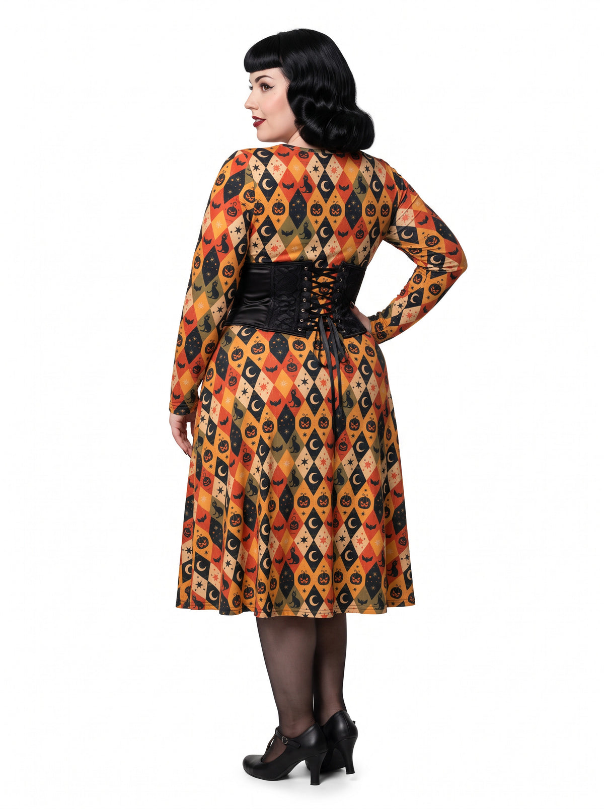 Plus-size model posing in a 1950s-style orange and black harlequin-patterned pinup midi swing dress featuring printed spooky pumpkins, bats, and crescent moons. The vintage-style dress shows a long-sleeve, fit-and-flare silhouette made from a smooth stretch fabric tailored for an inclusive, curvy fit.