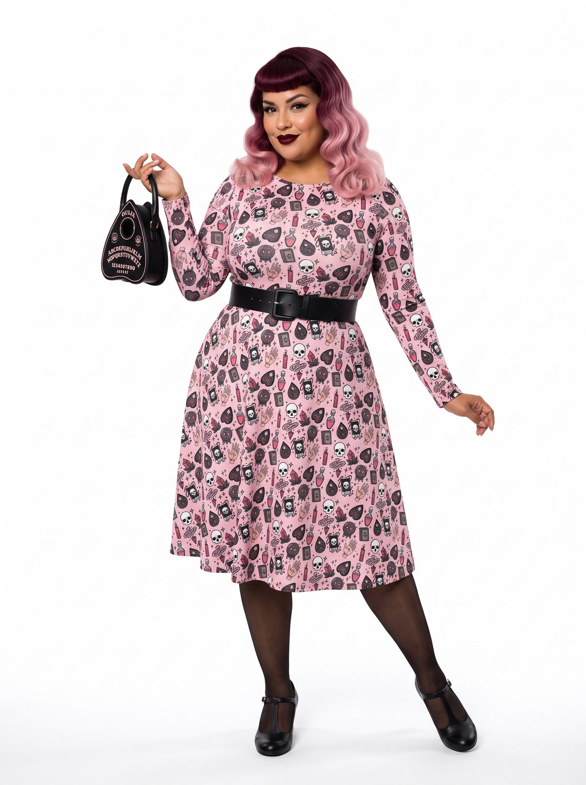 Plus-size model with a burgundy and pink ombre Bettie Page hairstyle posing in a 1950s pink witchy swing dress featuring skulls, planchettes, and tarot illustrations. The long-sleeve, fit-and-flare midi dress is showcasing an inclusive curvy fit on a white background.