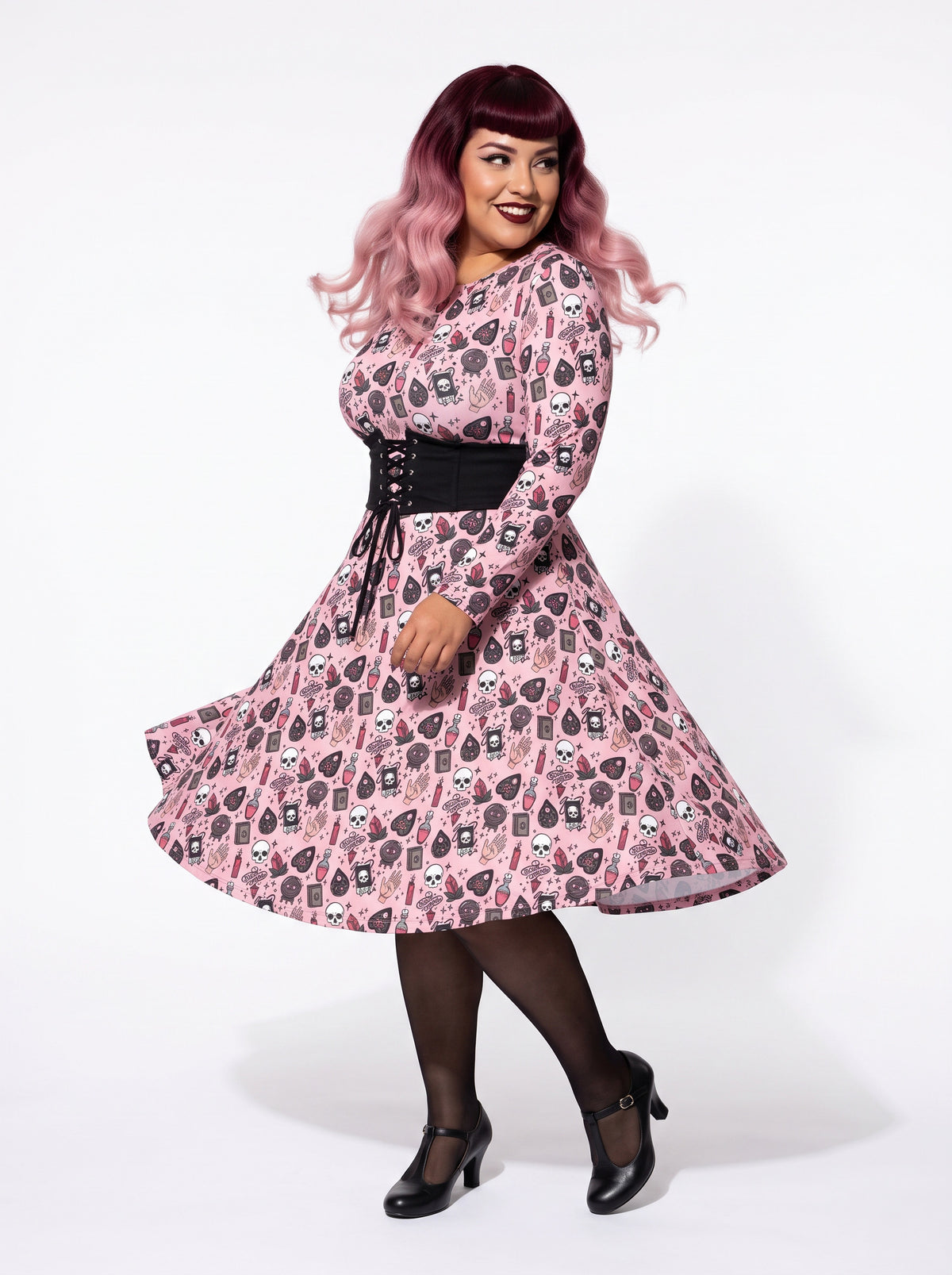 Plus-size model with a burgundy and pink ombre Bettie Page hairstyle posing in a 1950s pink witchy swing dress featuring skulls, planchettes, and tarot illustrations. The long-sleeve, fit-and-flare midi dress is styled with a black waist belt, showcasing an inclusive curvy fit on a white background.