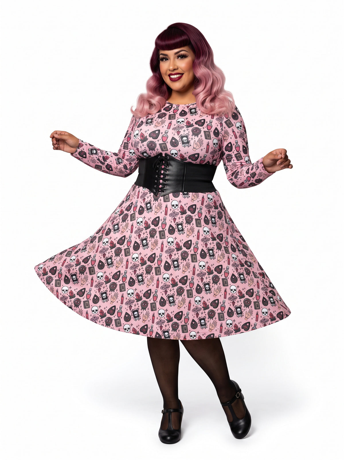 Plus-size model with a burgundy and pink ombre Bettie Page hairstyle posing in a 1950s pink witchy swing dress featuring skulls, planchettes, and tarot illustrations. The long-sleeve, fit-and-flare midi dress is showcasing an inclusive curvy fit on a white background.