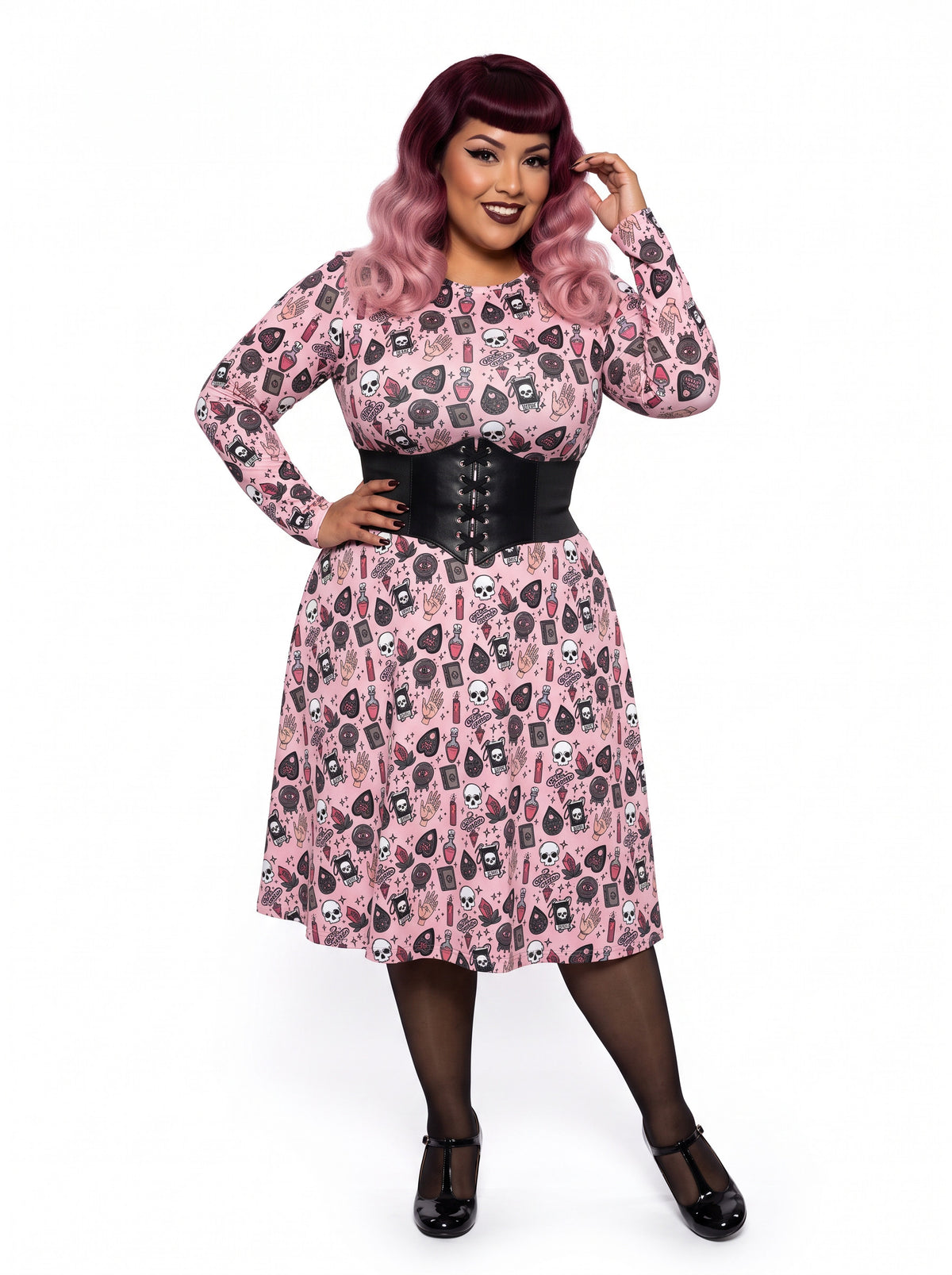 Plus-size model with a burgundy and pink ombre Bettie Page hairstyle posing in a 1950s pink witchy swing dress featuring skulls, planchettes, and tarot illustrations. The long-sleeve, fit-and-flare midi dress is showcasing an inclusive curvy fit on a white background.