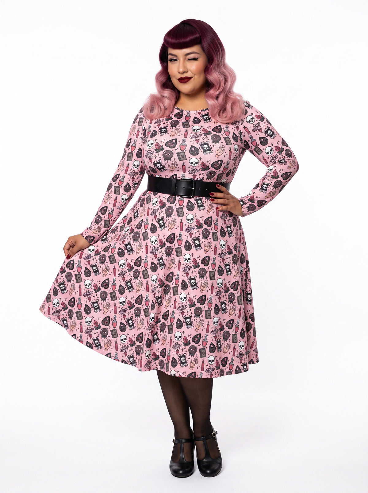 Plus-size model with a burgundy and pink ombre Bettie Page hairstyle posing in a 1950s pink witchy swing dress featuring skulls, planchettes, and tarot illustrations. The long-sleeve, fit-and-flare midi dress is showcasing an inclusive curvy fit on a white background.