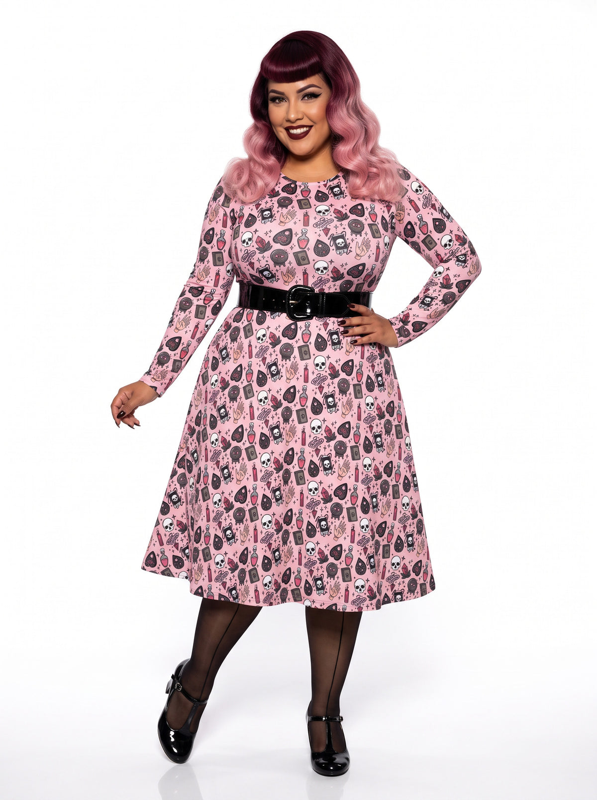 Plus-size model with a burgundy and pink ombre Bettie Page hairstyle posing in a 1950s pink witchy swing dress featuring skulls, planchettes, and tarot illustrations. The long-sleeve, fit-and-flare midi dress is showcasing an inclusive curvy fit on a white background.
