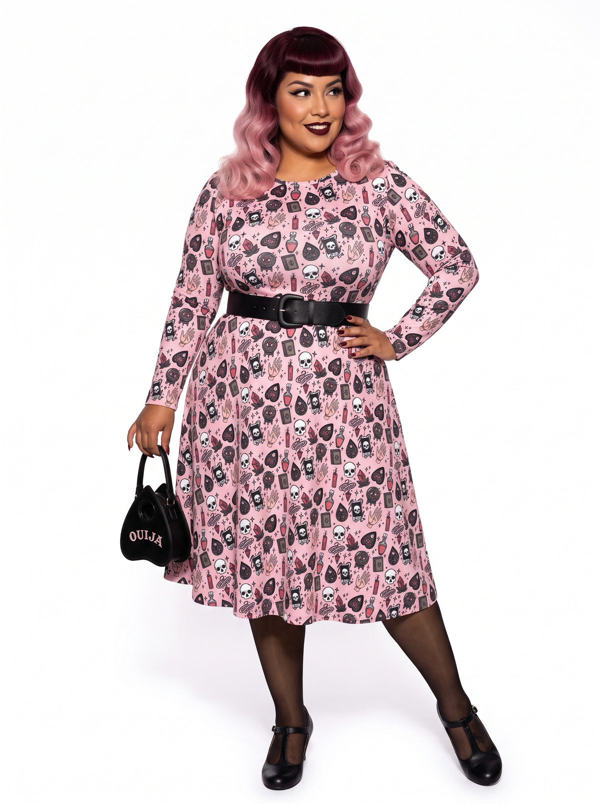 Plus-size model with a burgundy and pink ombre Bettie Page hairstyle posing in a 1950s pink witchy swing dress featuring skulls, planchettes, and tarot illustrations. The long-sleeve, fit-and-flare midi dress is showcasing an inclusive curvy fit on a white background.
