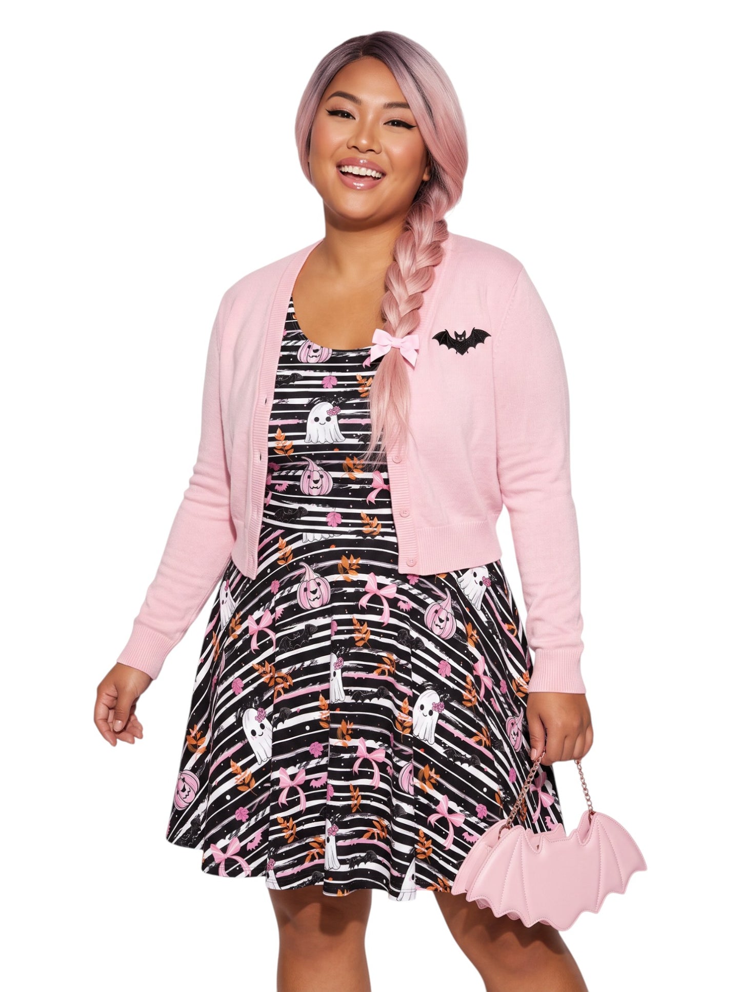 A smiling woman with braided pink hair showcases a sleeveless black-and-white striped Halloween skater dress featuring a cute goth print of ghosts with pink bows, bats, and pink jack-o'-lanterns. The dress has a flowy, fit-and-flare silhouette that highlights her hourglass shape.