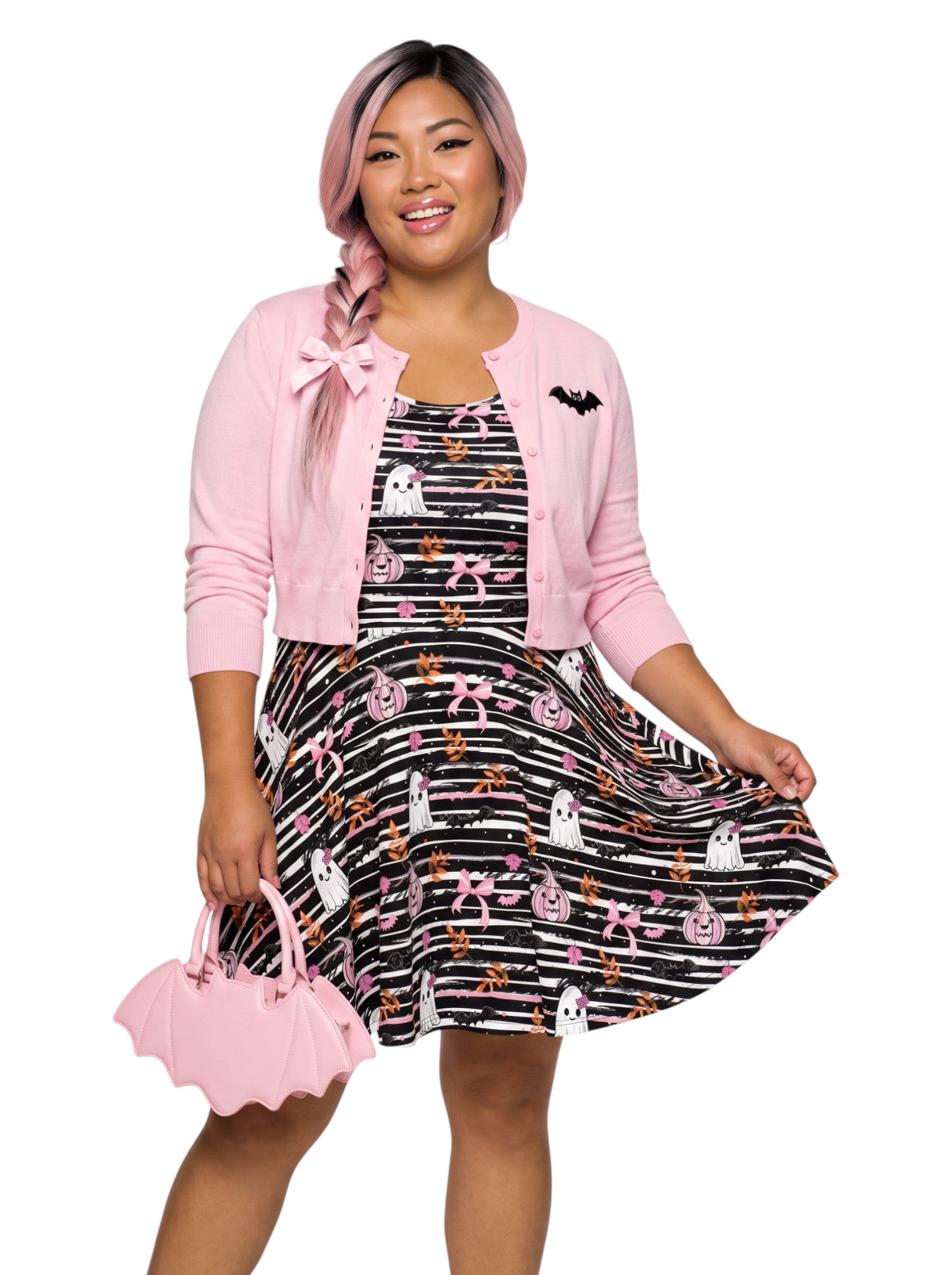 A smiling woman with braided pink hair showcases a sleeveless black-and-white striped Halloween skater dress featuring a cute goth print of ghosts with pink bows, bats, and pink jack-o'-lanterns. The dress has a flowy, fit-and-flare silhouette that highlights her hourglass shape.
