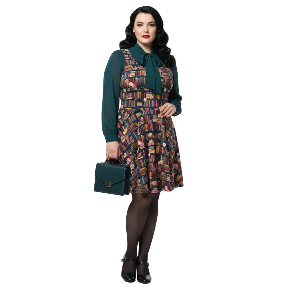 Plus size woman wearing a dark academia bookshelf print skater dress with a green pussy-bow blouse and matching handbag.
