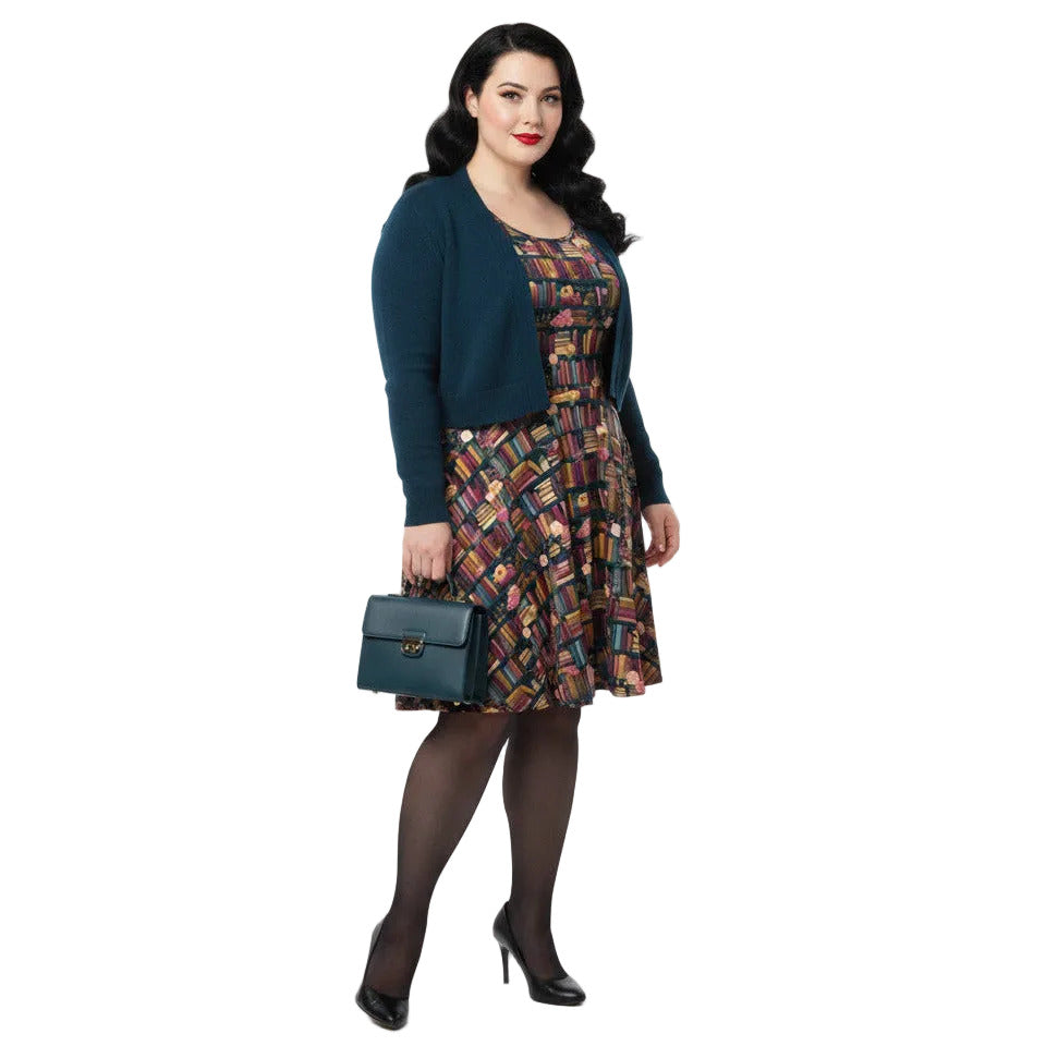 Plus size woman wearing a dark academia bookshelf print skater dress with a teal green cardigan sweater and matching handbag.
