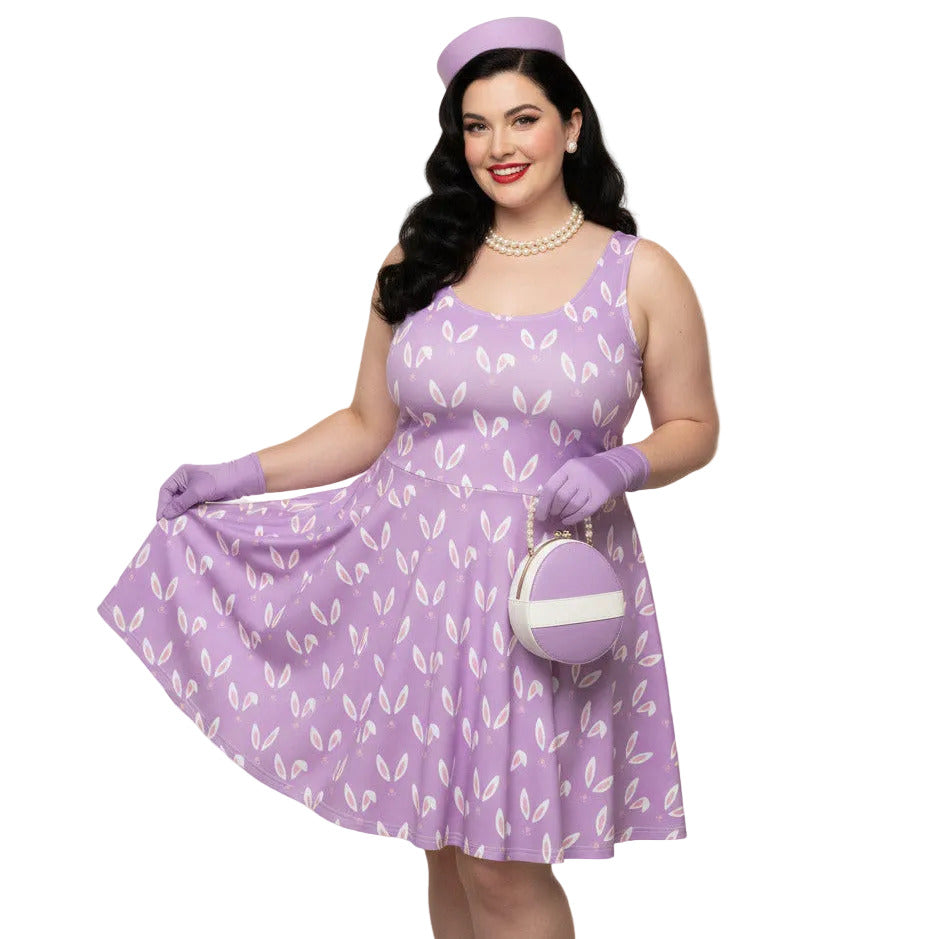 Curvy woman wearing a lavender purple plus size 1950s vintage style swing dress with white Easter bunny print, matching pillbox hat, and pearl necklace.