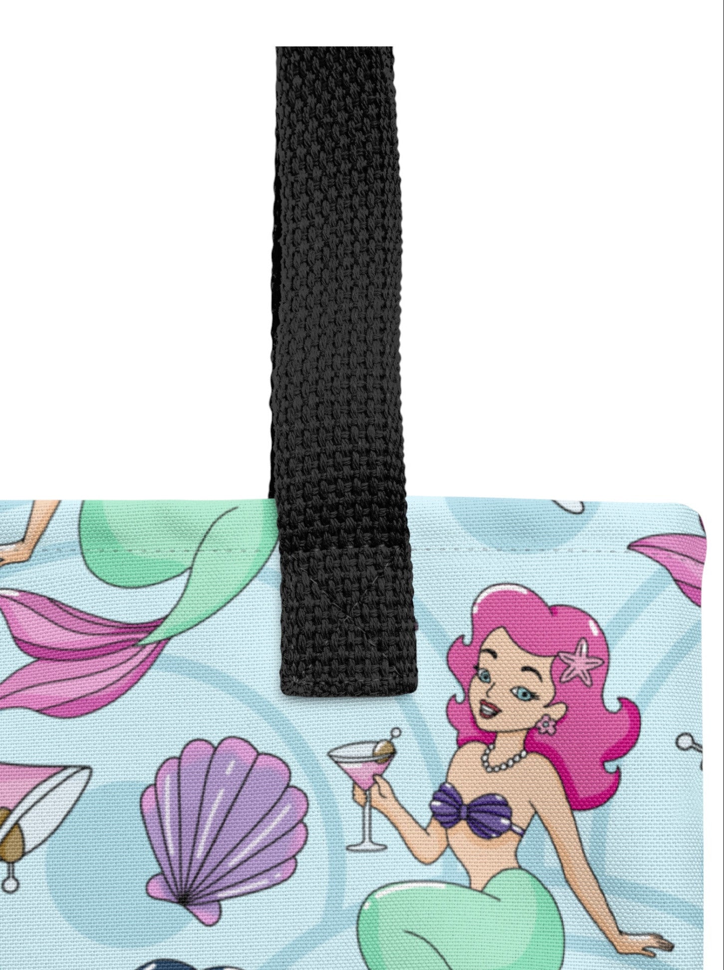 A close-up of a tote bag featuring a light blue mermaid-themed print with pink, purple, and green details, complete with a black woven handle.