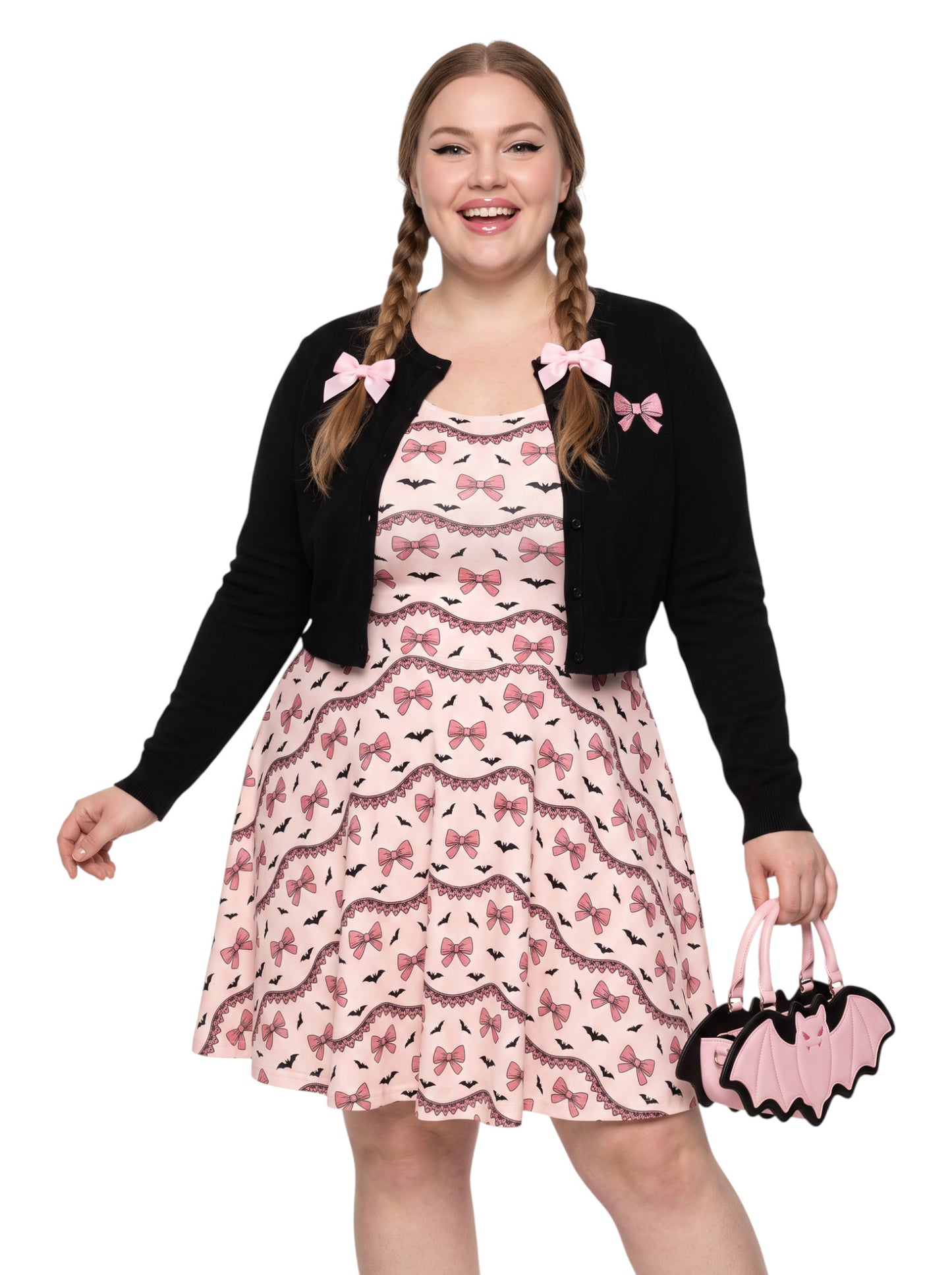 Plus size woman with braided hair wearing a pink sleeveless fit and flare pastel goth coquette dress with a pattern of black spooky bats, pink bows, and lace details.