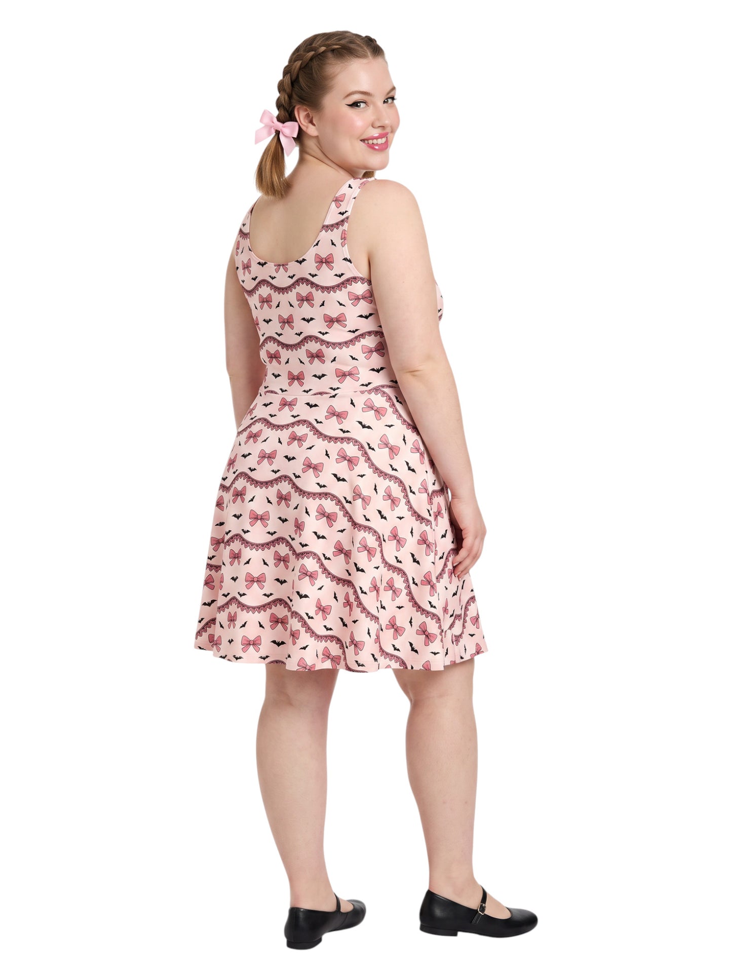 Plus size woman with braided hair wearing a pink sleeveless fit and flare pastel goth coquette dress with a pattern of black spooky bats, pink bows, and lace details.