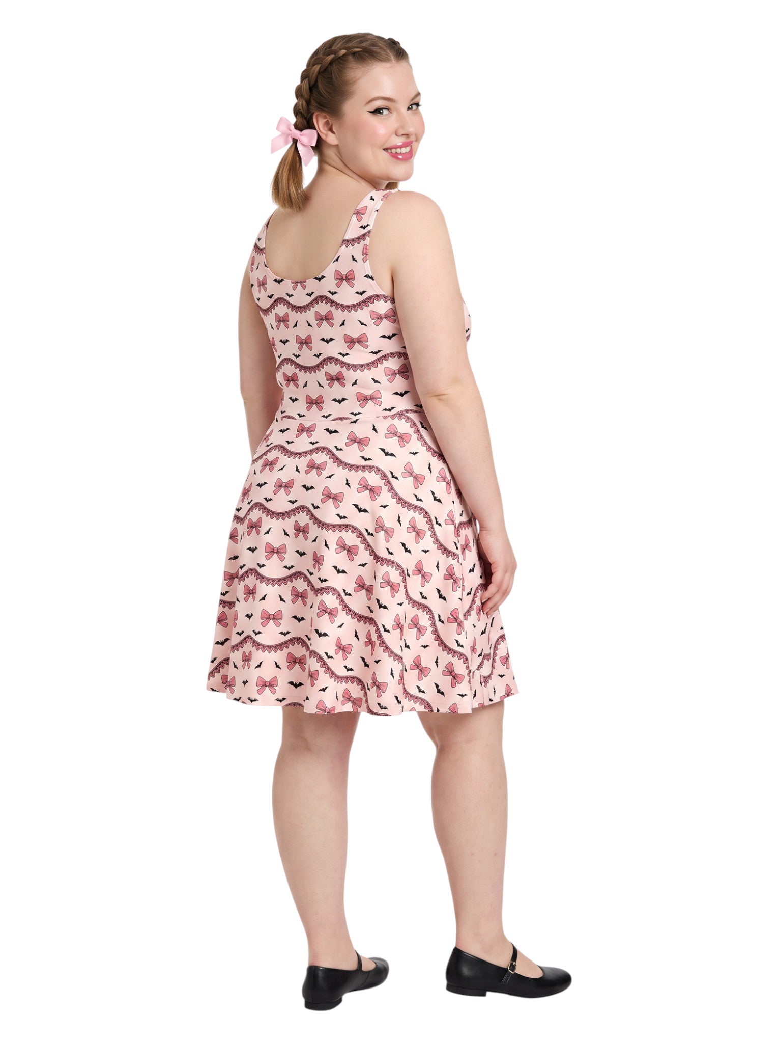 Plus size woman with braided hair wearing a pink sleeveless fit and flare pastel goth coquette dress with a pattern of black spooky bats, pink bows, and lace details.
