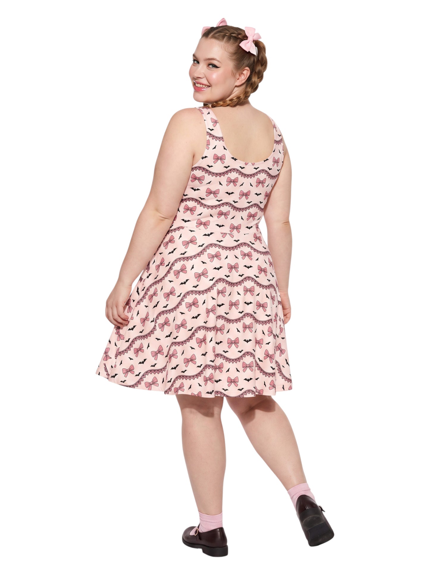 Plus size woman with braided hair wearing a pink sleeveless fit and flare pastel goth coquette dress with a pattern of black spooky bats, pink bows, and lace details.