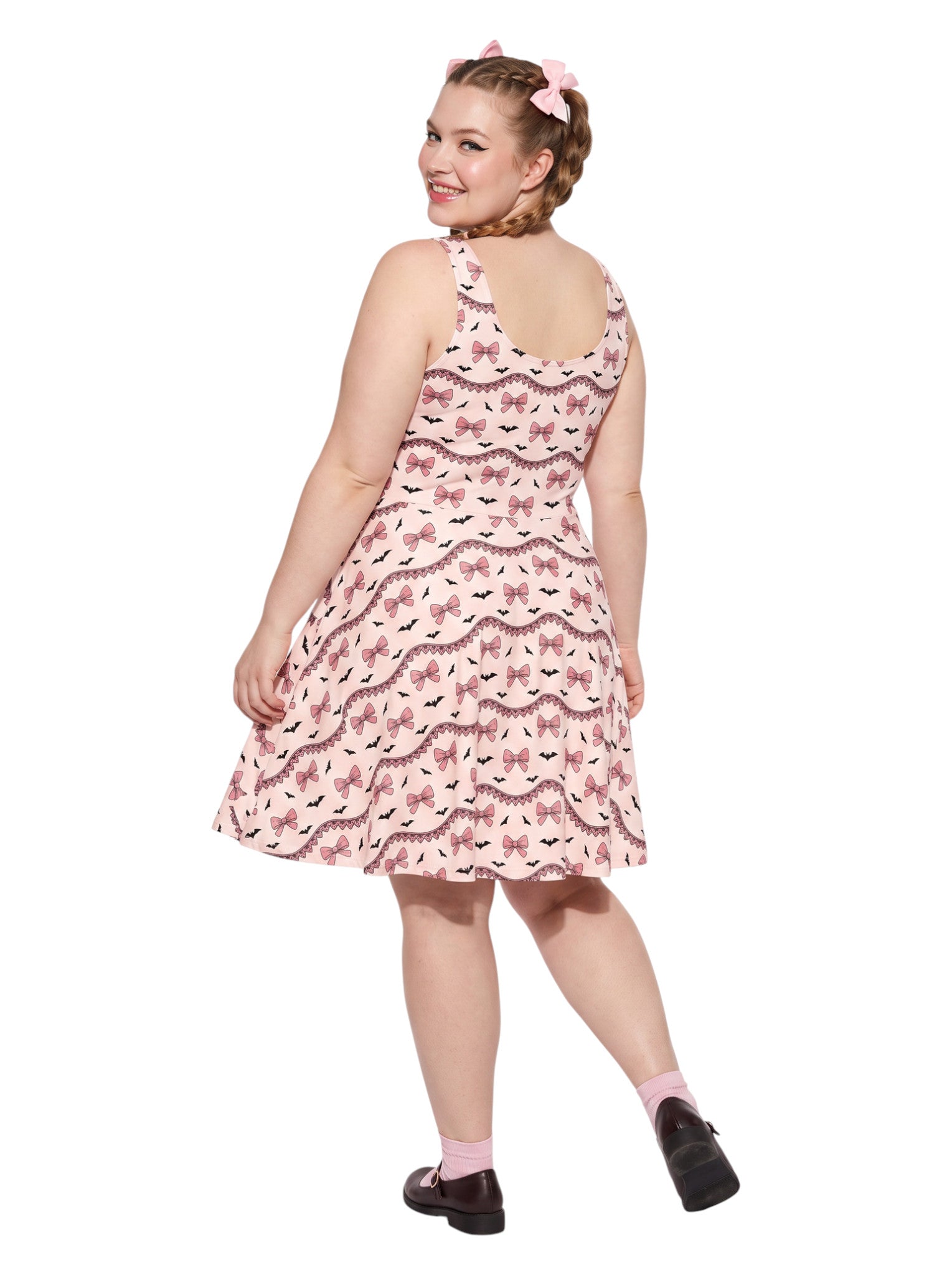 Plus size woman with braided hair wearing a pink sleeveless fit and flare pastel goth coquette dress with a pattern of black spooky bats, pink bows, and lace details.