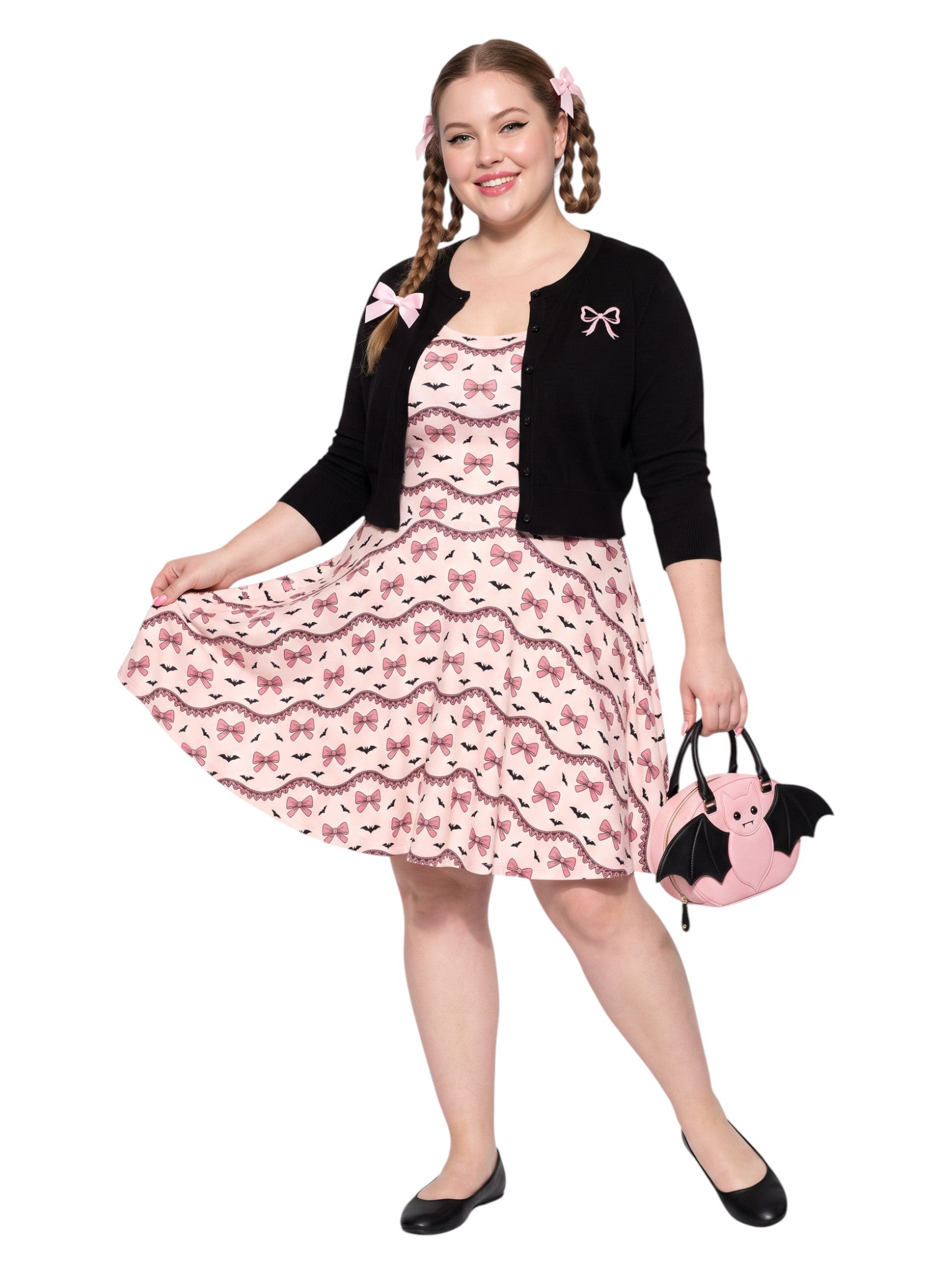 Plus size woman with braided hair wearing a pink sleeveless fit and flare pastel goth coquette dress with a pattern of black spooky bats, pink bows, and lace details.