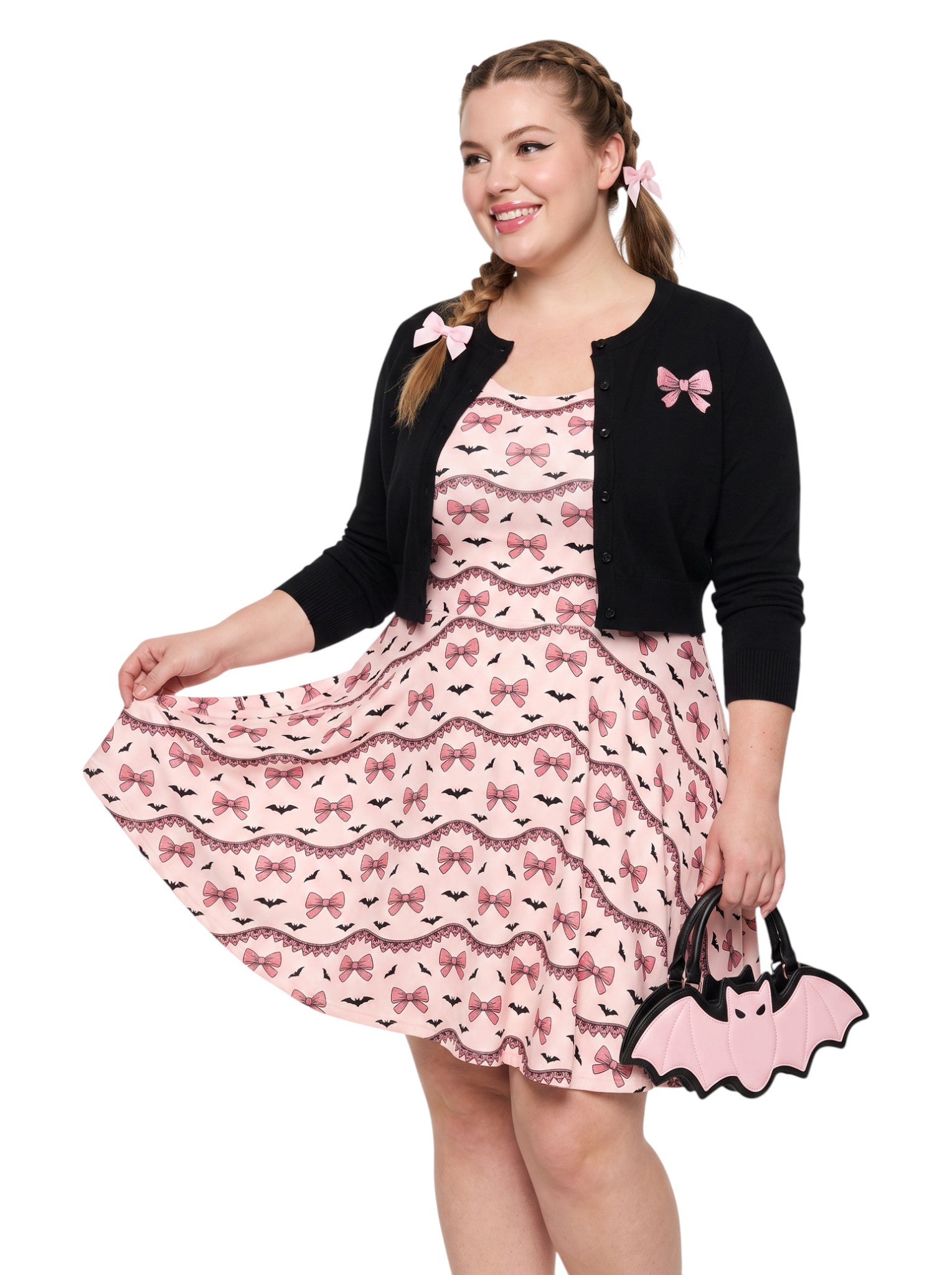 Plus size woman with braided hair wearing a pink sleeveless fit and flare pastel goth coquette dress with a pattern of black spooky bats, pink bows, and lace details.