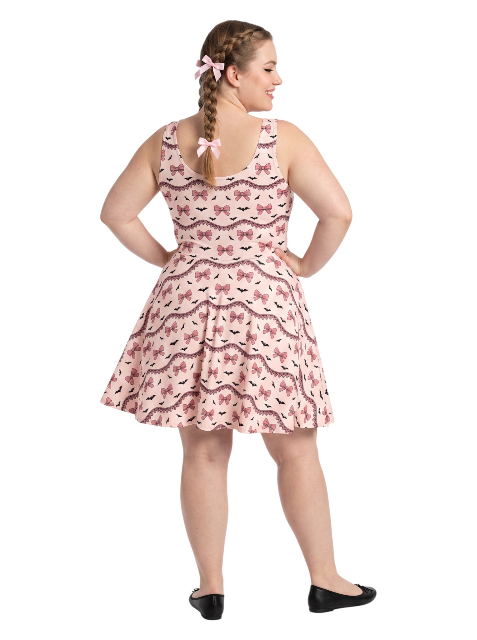 Plus size woman with braided hair wearing a pink sleeveless fit and flare pastel goth coquette dress with a pattern of black spooky bats, pink bows, and lace details.