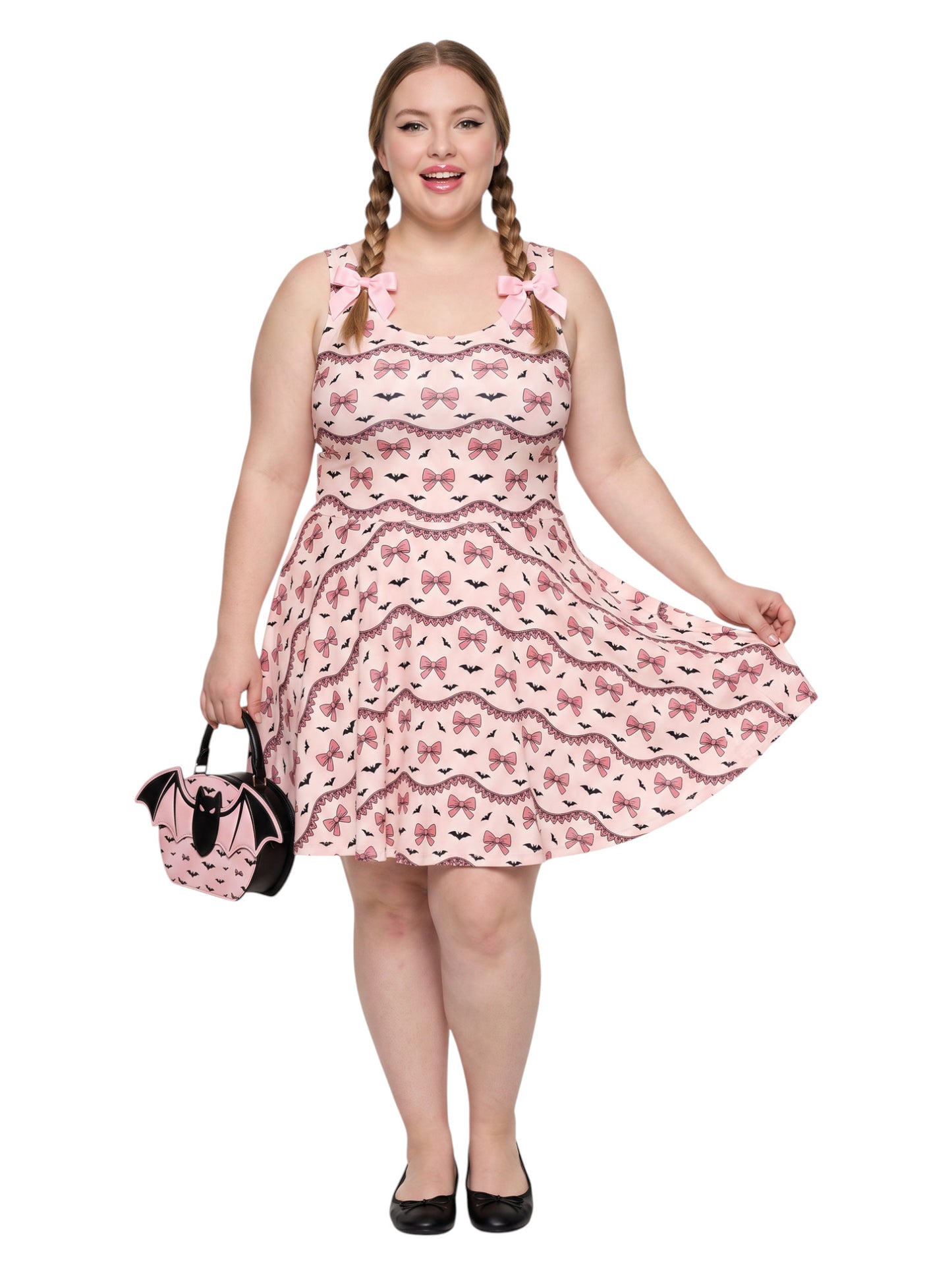 Plus size woman with braided hair wearing a pink sleeveless fit and flare pastel goth coquette dress with a pattern of black spooky bats, pink bows, and lace details.