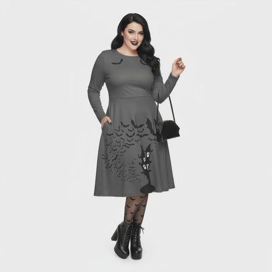 Plus size woman wearing a grey long sleeve goth pinup dress with black bat and haunted house print featuring functional side pockets.