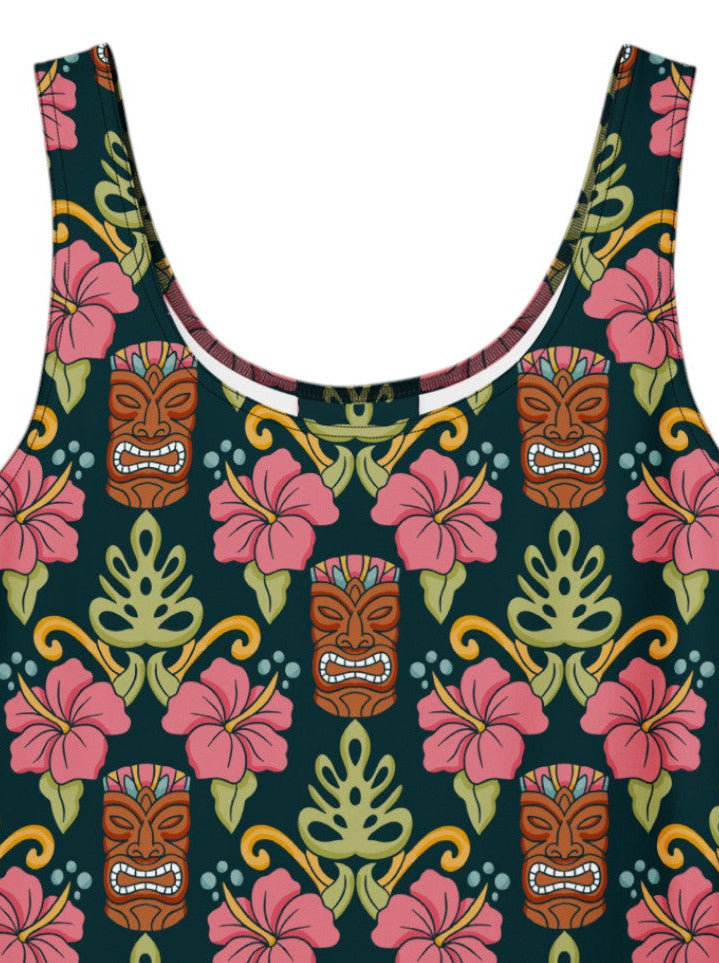 A dark green 1950s-style pinup rockabilly swing dress with an all-over tropical print of tiki masks, pink hibiscus flowers, and monstera leaves. 