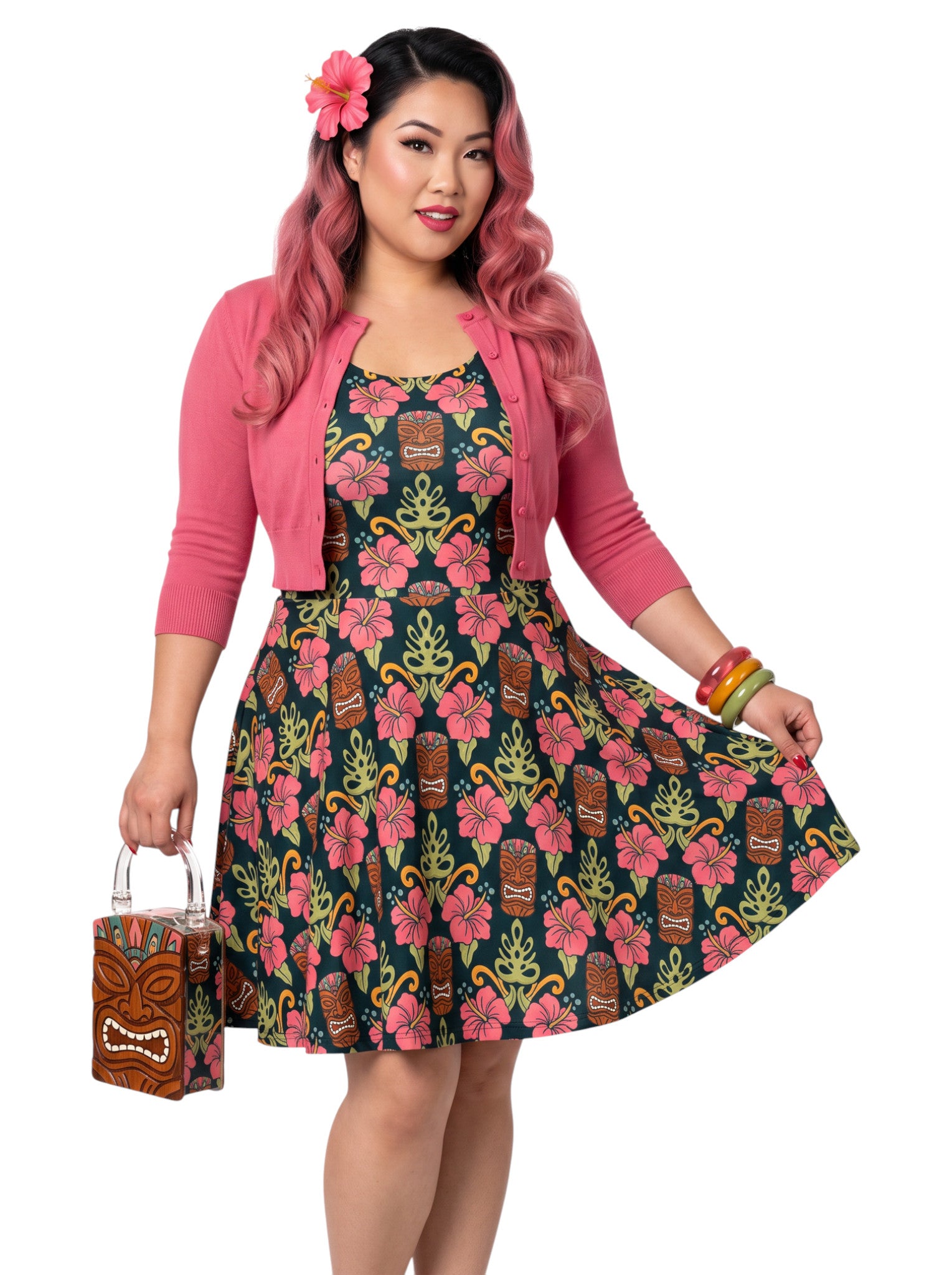 A plus size woman wearing a dark green 1950s-style pinup rockabilly swing dress with an all-over tropical print of tiki masks, pink hibiscus flowers, and monstera leaves. 
