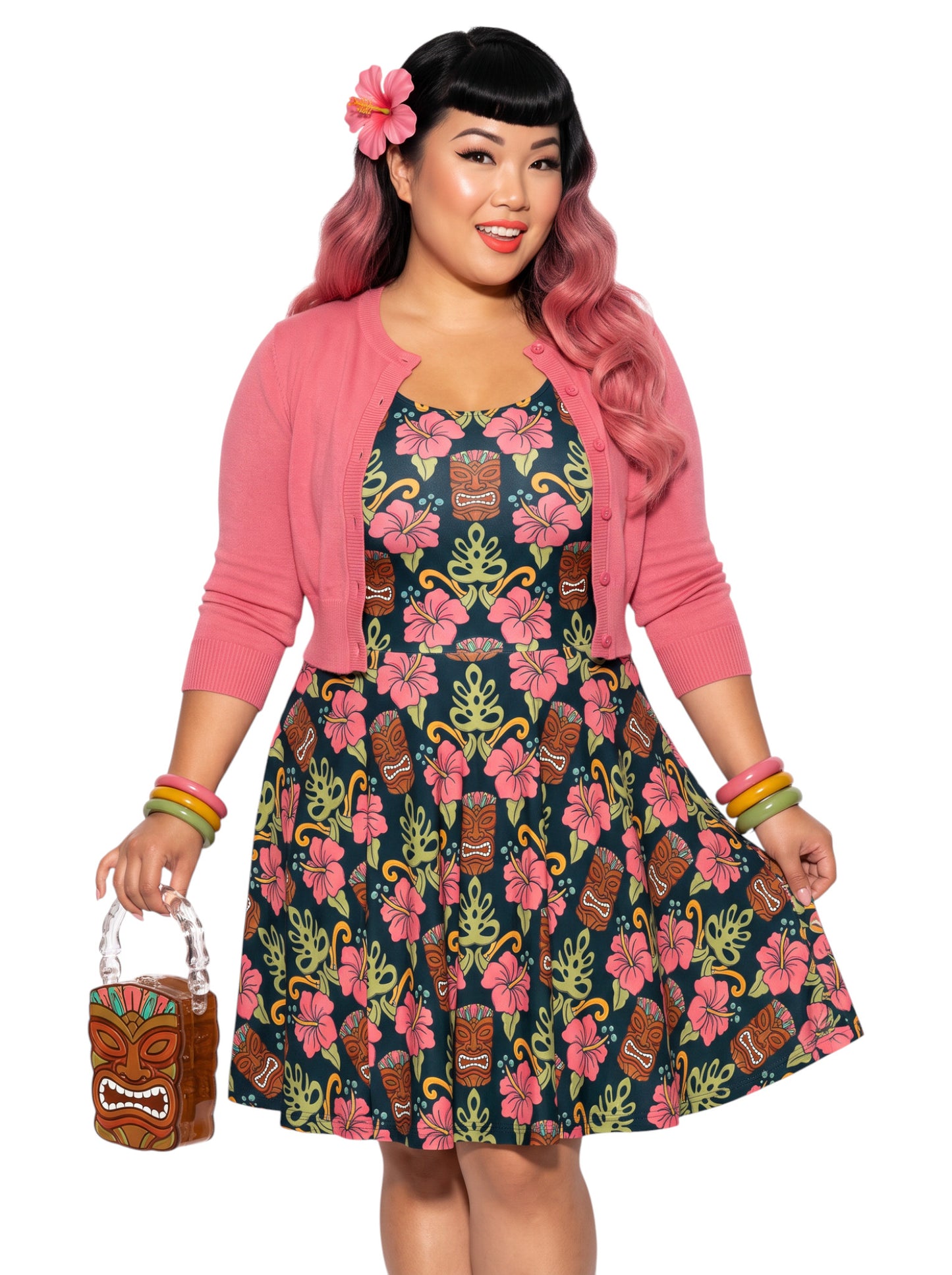 A plus size woman wearing a dark green 1950s-style pinup rockabilly swing dress with an all-over tropical print of tiki masks, pink hibiscus flowers, and monstera leaves. 