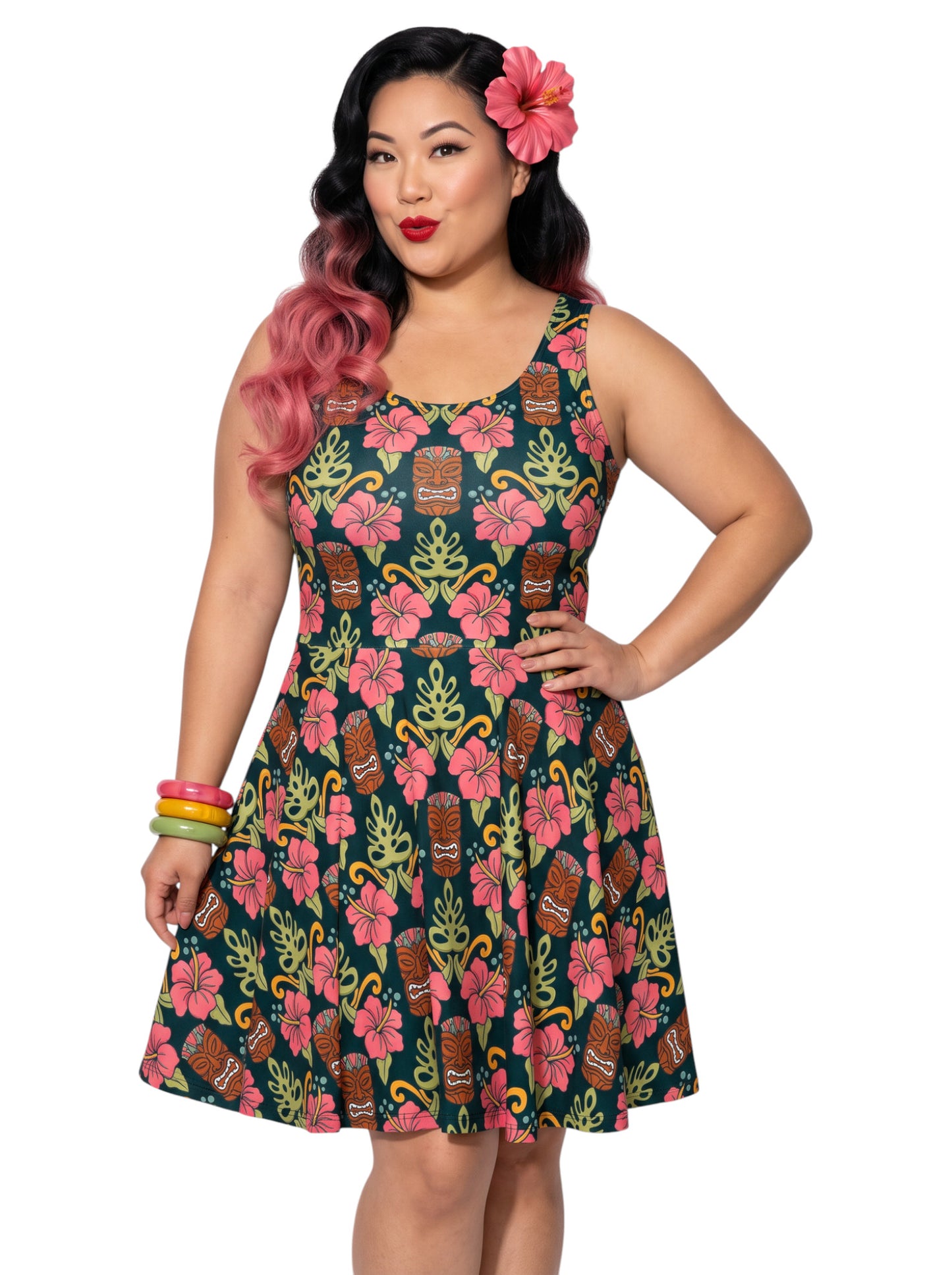 A plus size woman wearing a dark green 1950s-style pinup rockabilly swing dress with an all-over tropical print of tiki masks, pink hibiscus flowers, and monstera leaves. 