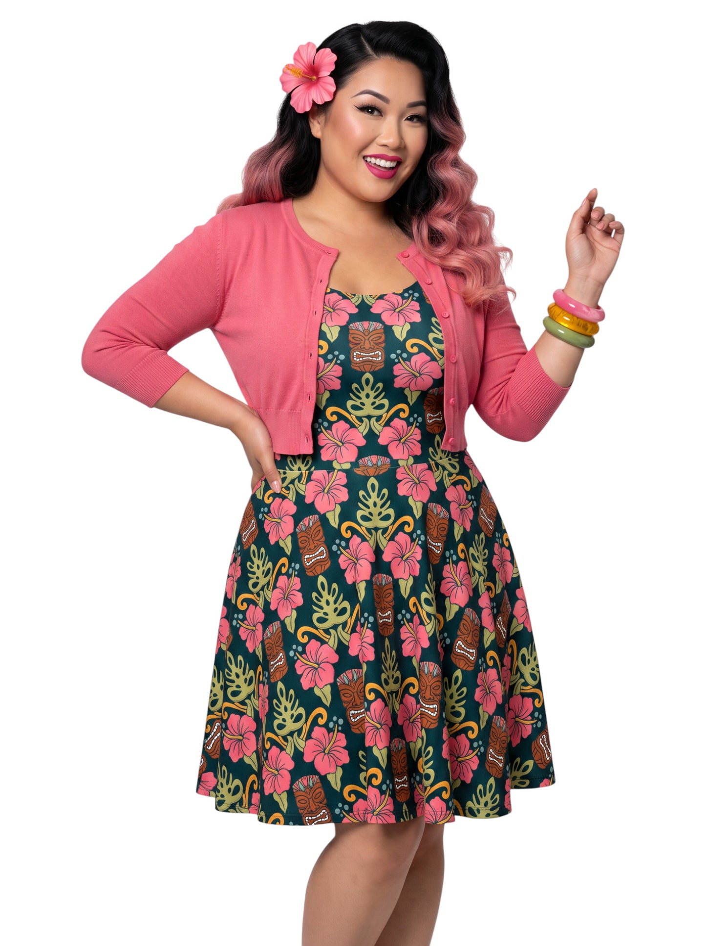 A plus size woman wearing a dark green 1950s-style pinup rockabilly swing dress with an all-over tropical print of tiki masks, pink hibiscus flowers, and monstera leaves. 