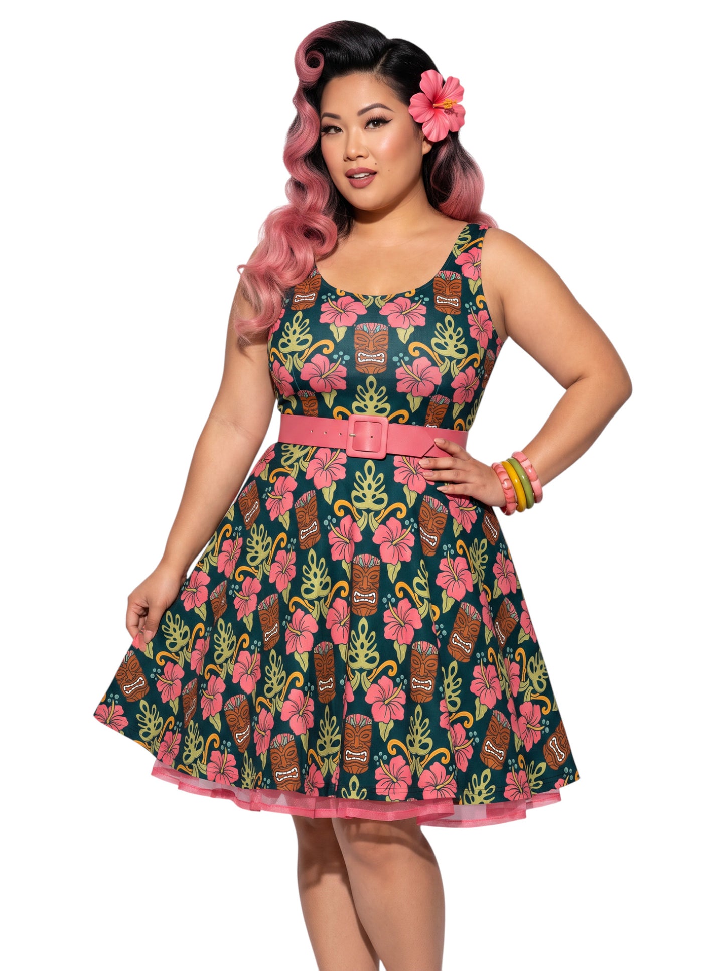 A plus size woman wearing a dark green 1950s-style pinup rockabilly swing dress with an all-over tropical print of tiki masks, pink hibiscus flowers, and monstera leaves. 