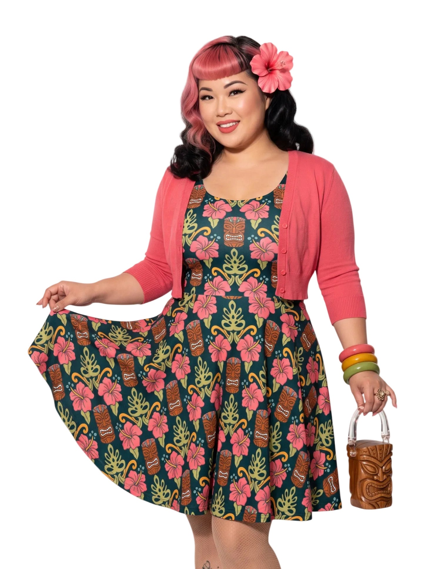 A plus size woman wearing a dark green 1950s-style pinup rockabilly swing dress with an all-over tropical print of tiki masks, pink hibiscus flowers, and monstera leaves. 